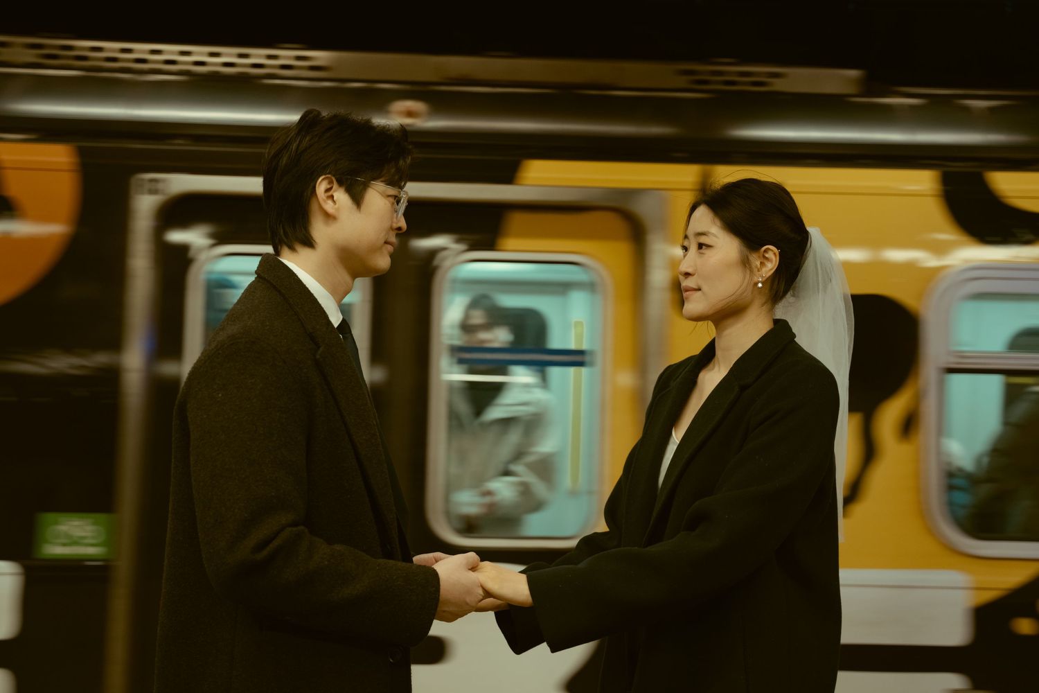 Two people in black formal attire hold hands on a subway platform with a yellow train passing behind them.