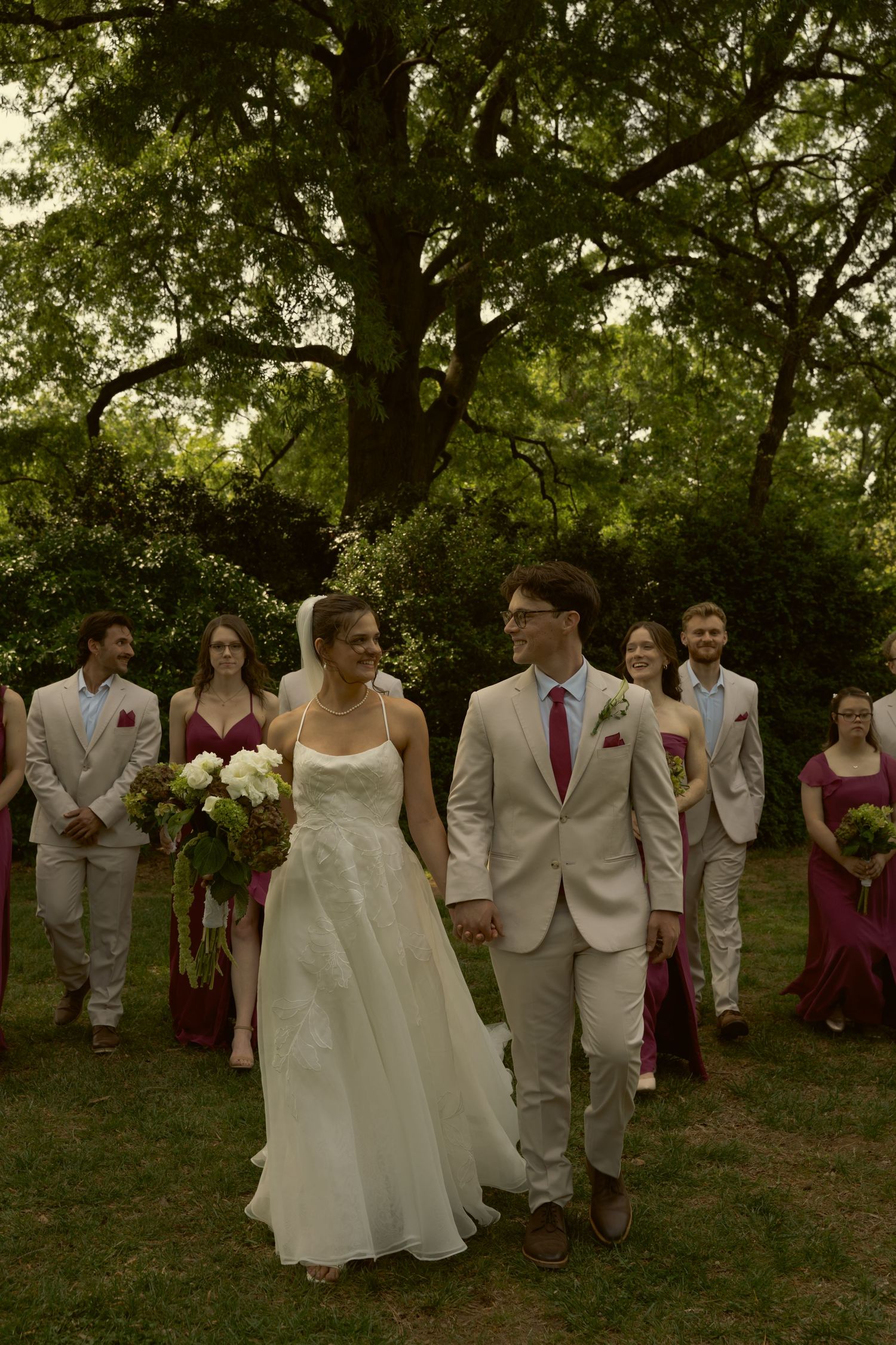 Newlyweds walk hand-in-hand with wedding party in burgundy and tan attire outdoors under lush green trees.