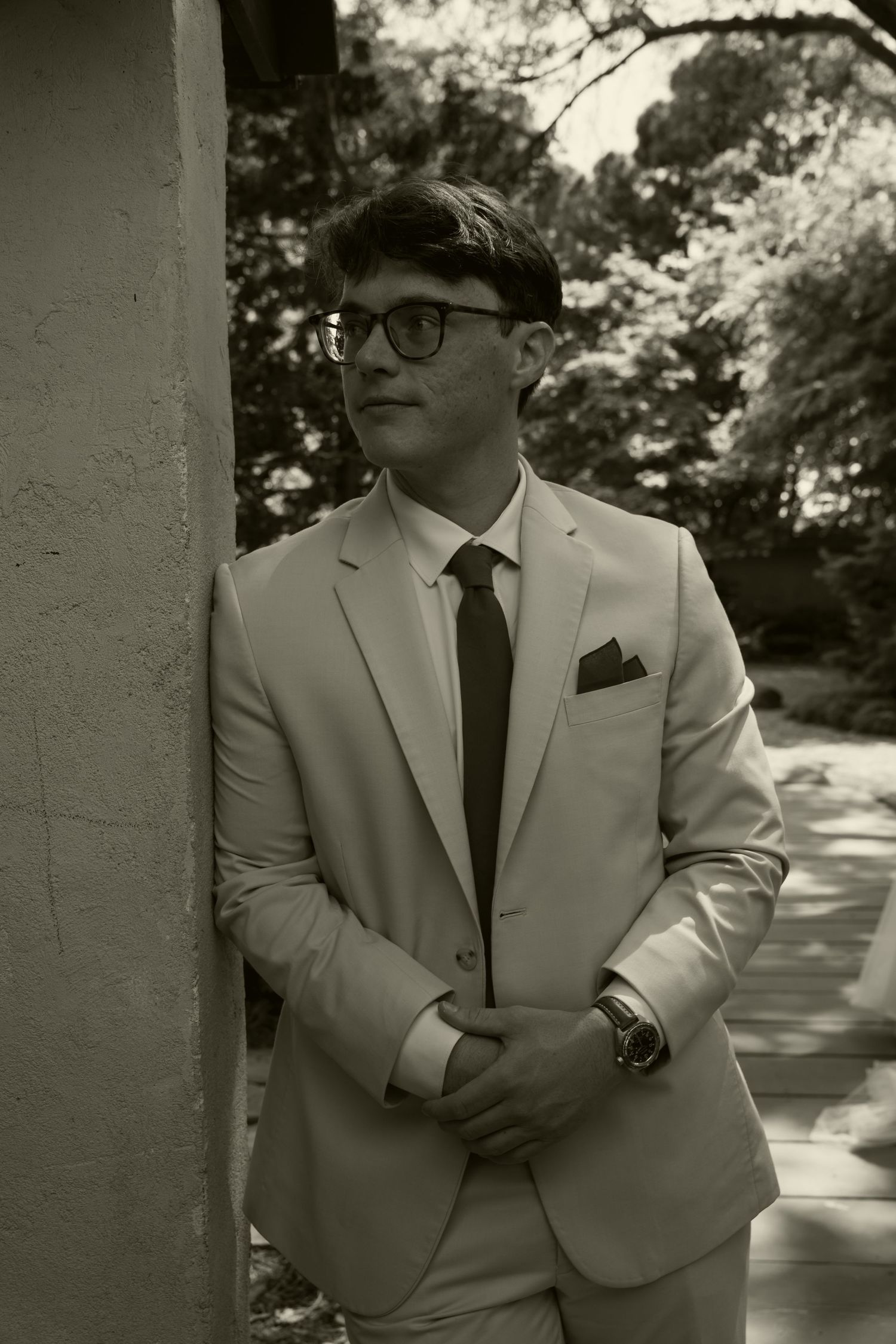 Black and white portrait of groom in tan suit and glasses leaning against a wall outdoors.