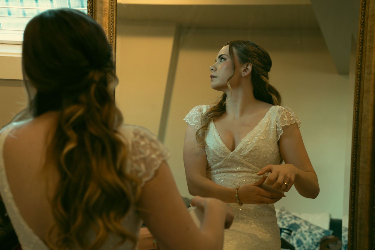A bride in a lace wedding dress looks at her reflection in a mirror during wedding preparations.
