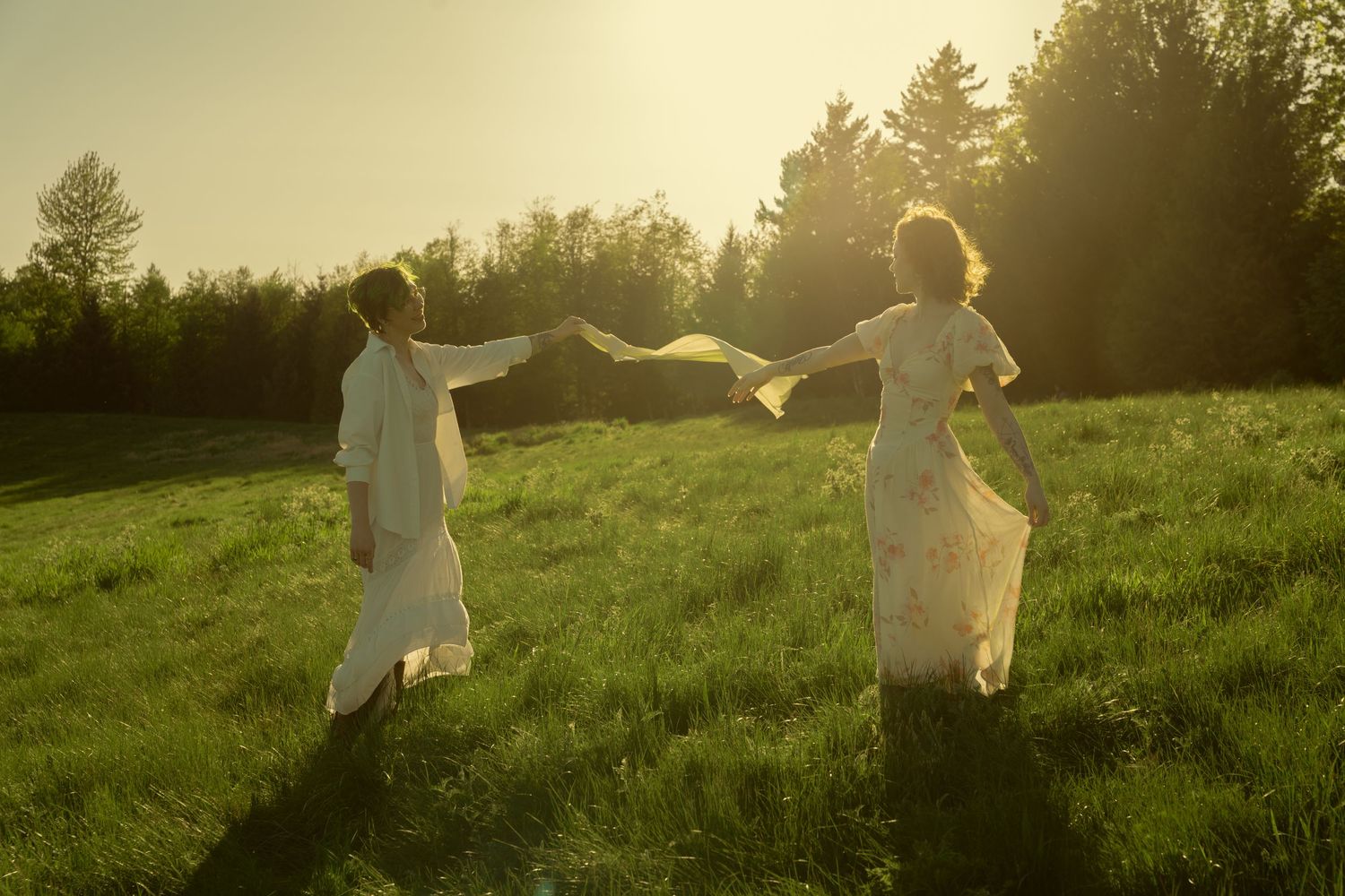 Two people in white flowing outfits hold hands running through a sunlit green meadow at golden hour.