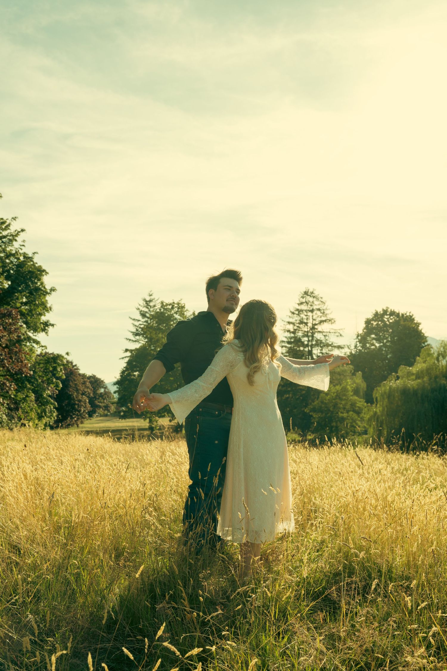 A couple embraces in a sunlit golden field, the woman in white dress and man in dark outfit, surrounded by lush green trees.