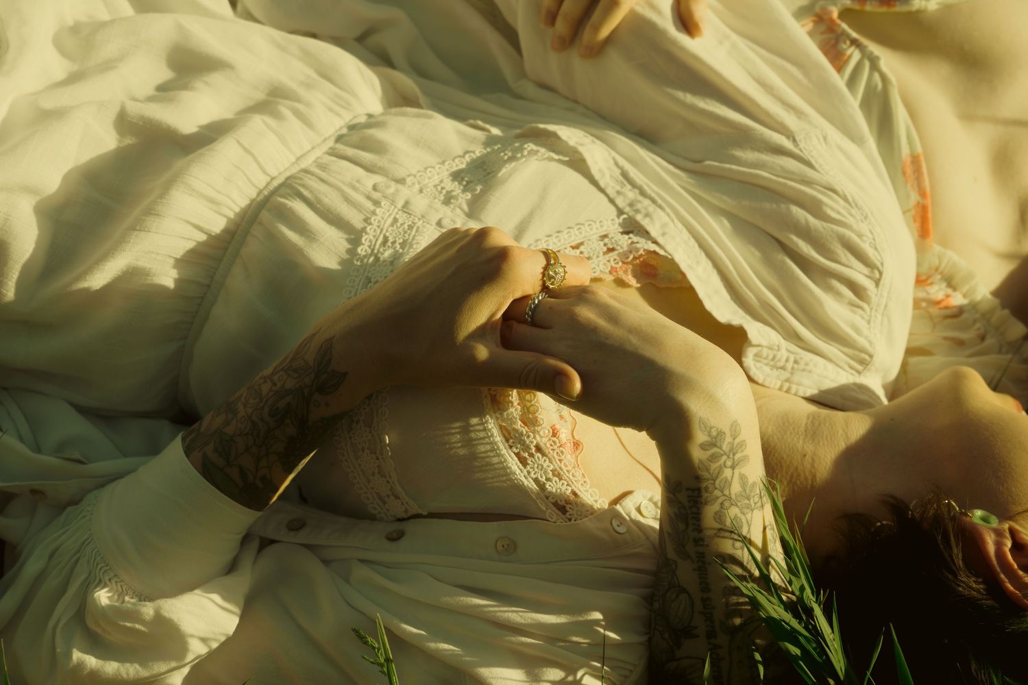 Person in white dress lying in grass, hands clasped showing tattoos and ring in warm golden sunlight.