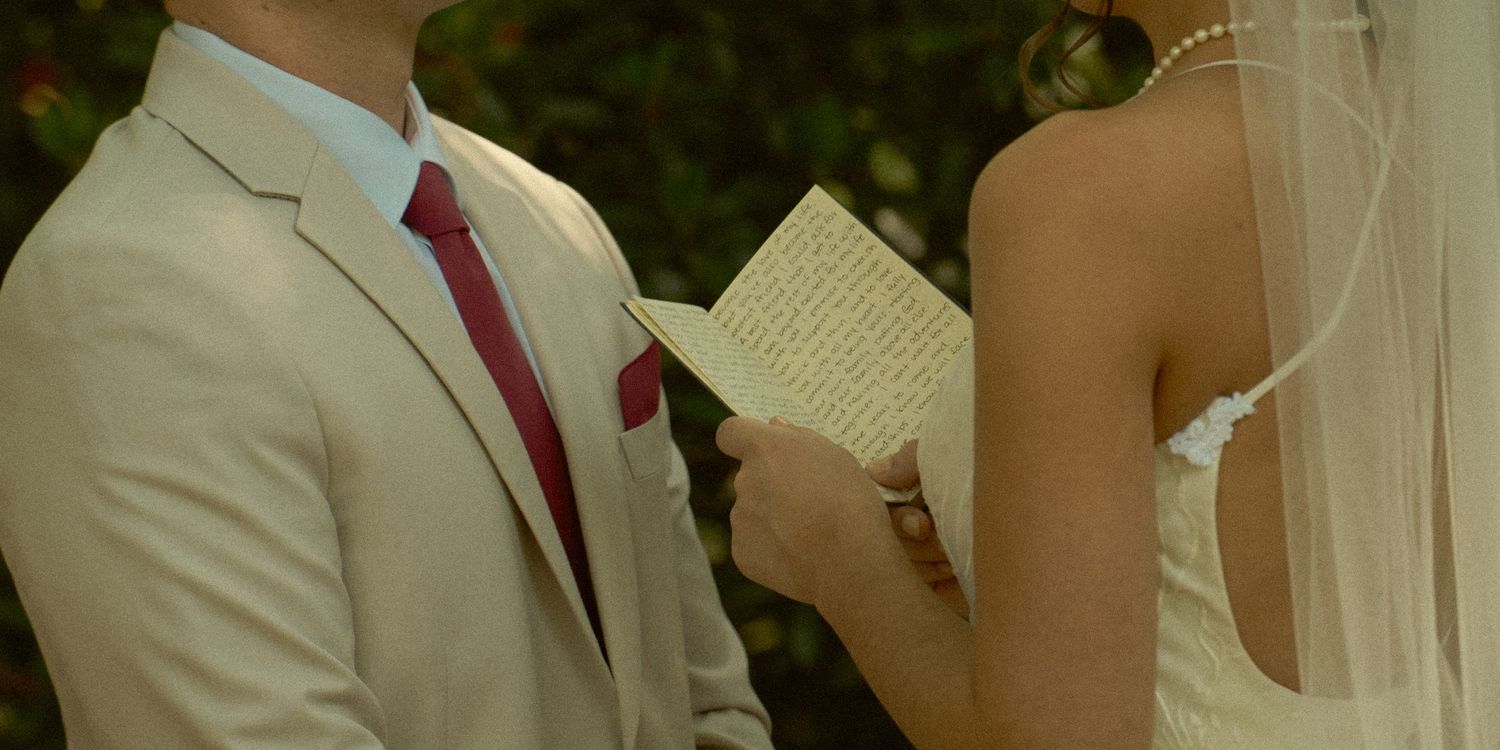 Groom in beige suit with red tie reads wedding vows from paper to bride in white dress and veil outdoors.