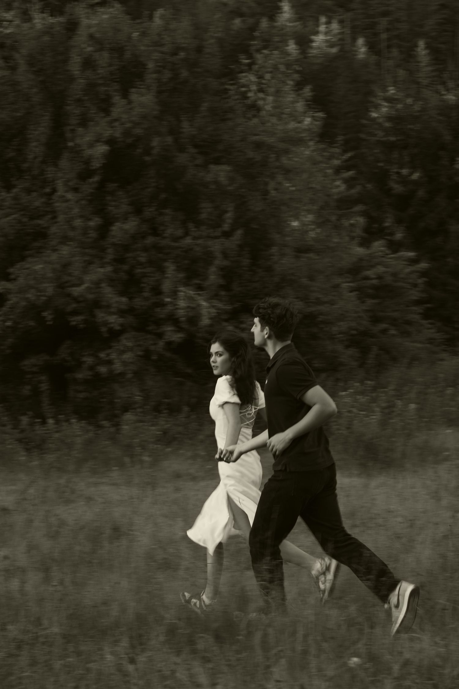 Black and white photo of a couple running through a grassy field with trees in the background.