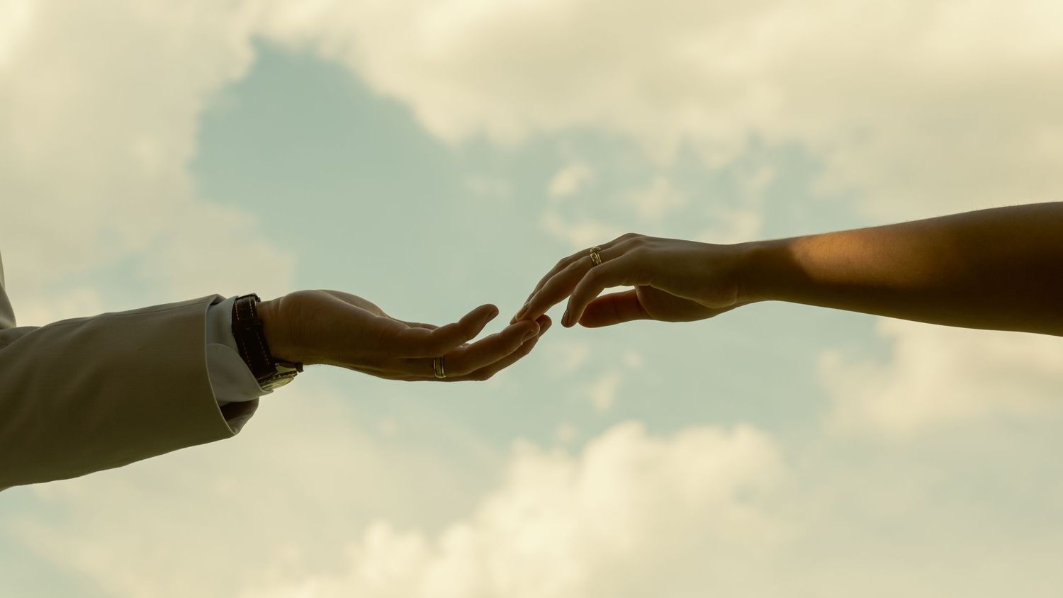 Two hands reaching toward each other against a cloudy sky, symbolizing connection and unity.