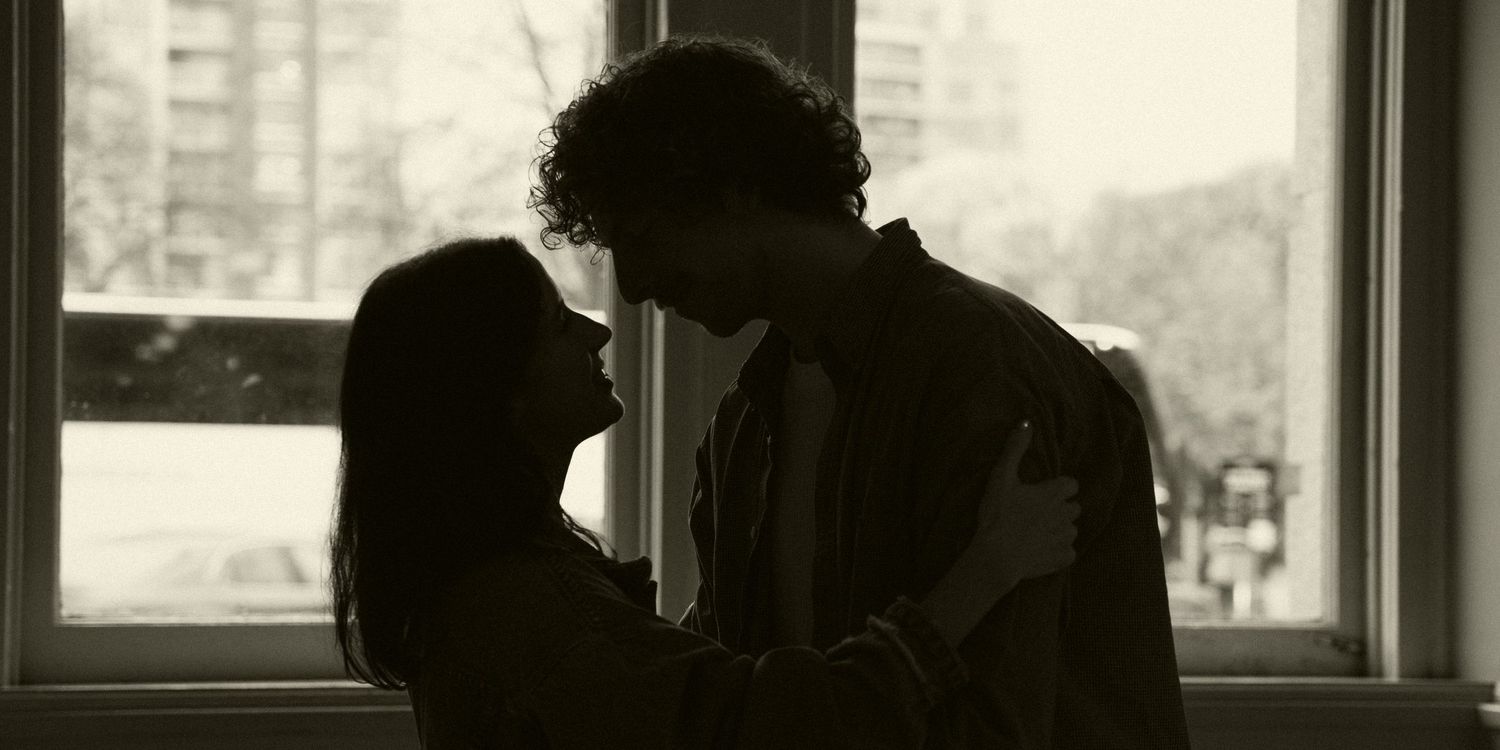 A couple stands in silhouette against a bright window in a dimly lit room.