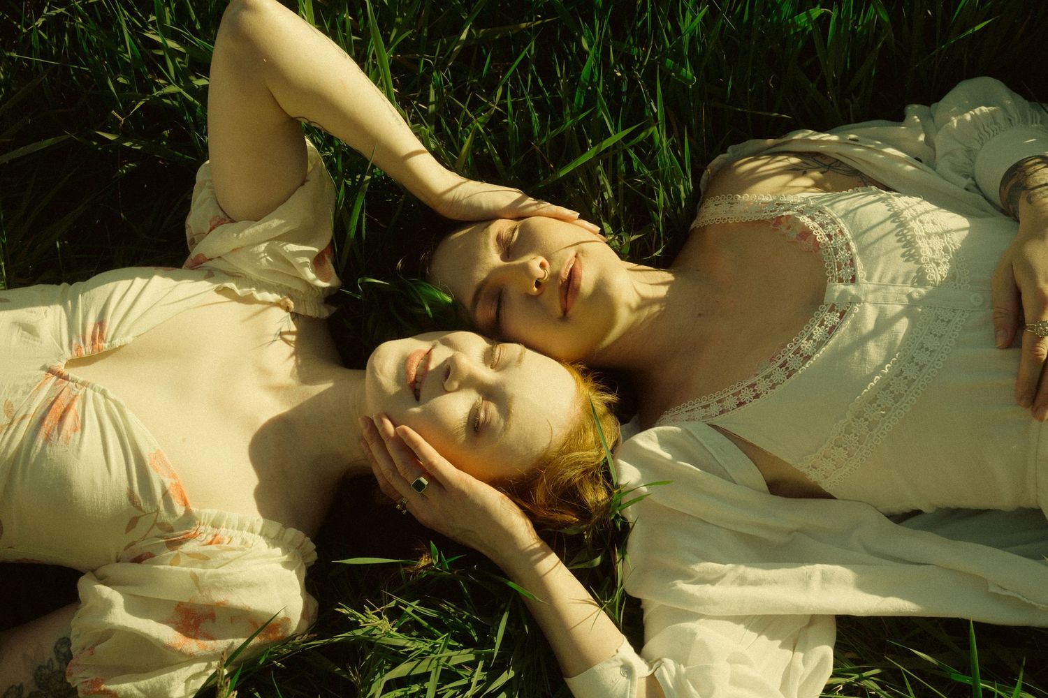 Two women in vintage cream dresses resting peacefully on green grass in warm sunlight.