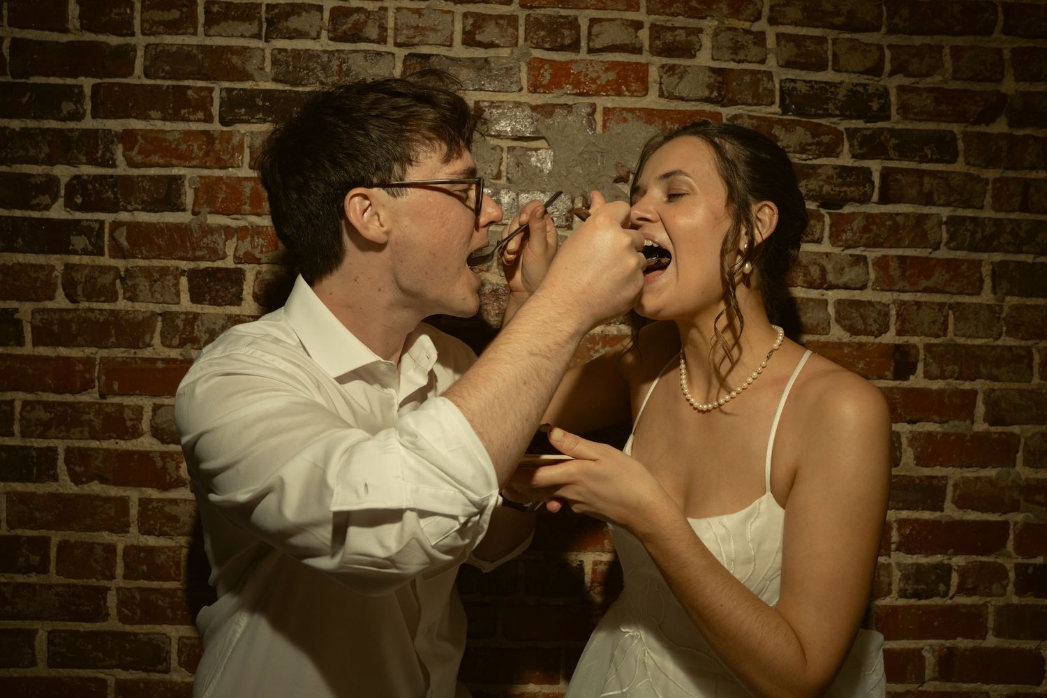 Bride and groom sharing a playful moment feeding each other at their wedding reception near a brick wall.