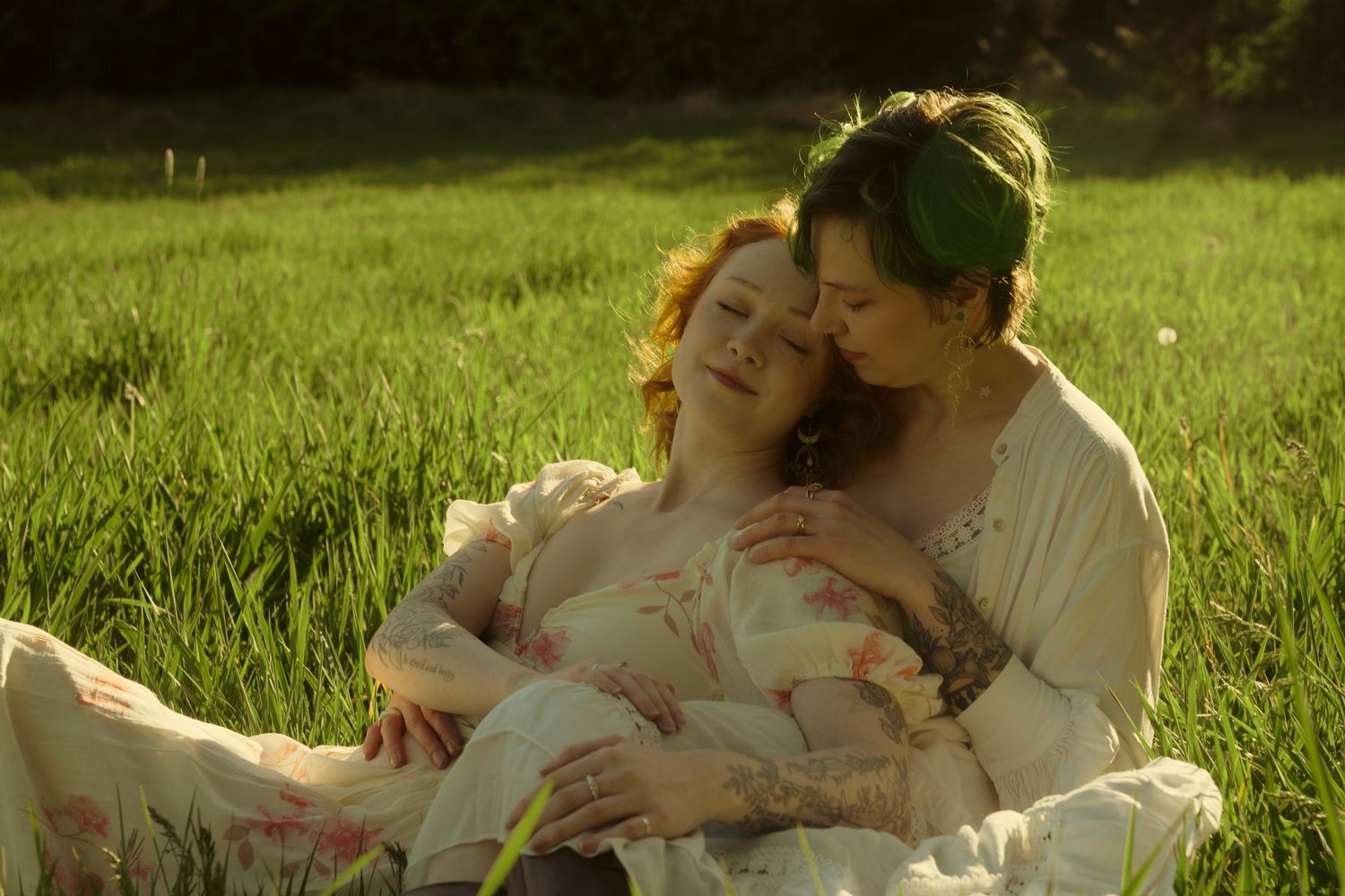 Two women cuddle lovingly in a lush green field, bathed in warm golden afternoon sunlight.