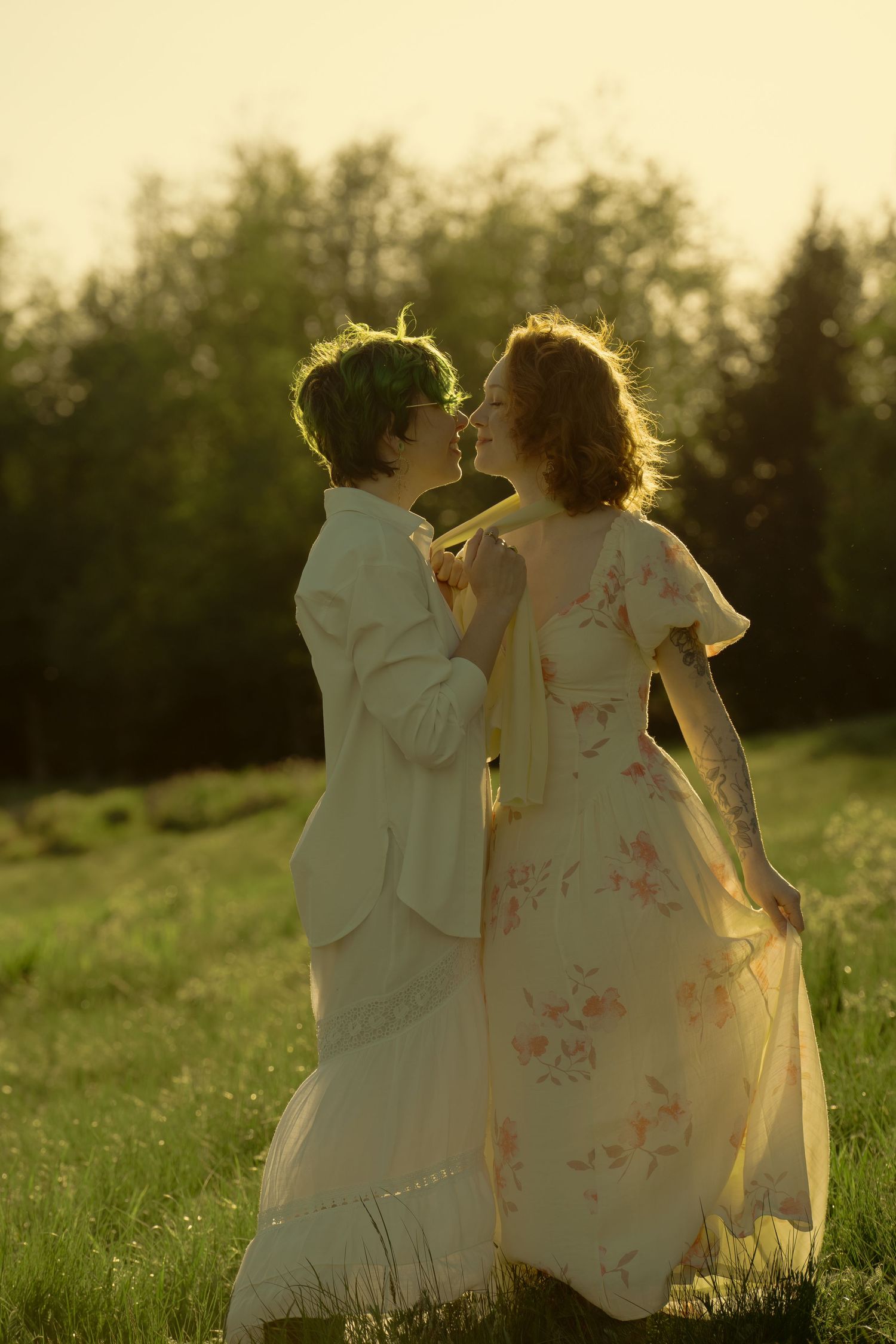 Two women in elegant dresses share a romantic moment in a sunlit meadow at golden hour.