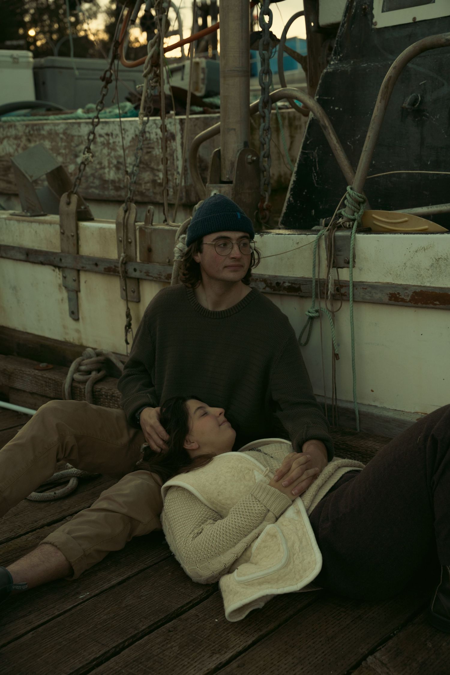 Two young people relaxing on a wooden boat deck at dusk, woman resting her head on man's lap.