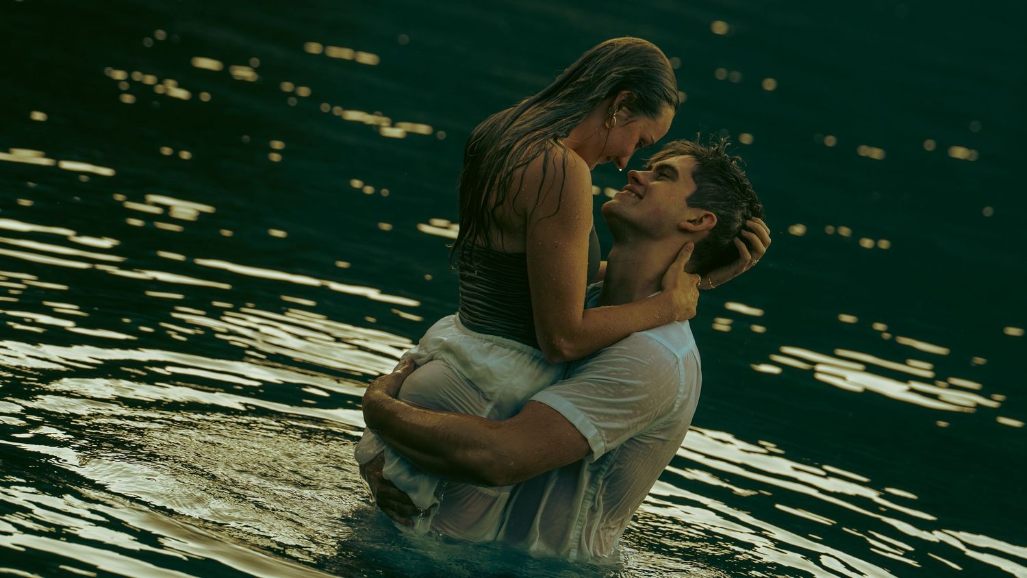 A romantic couple embraces in a shimmering lake, the man lifting the woman as they share an intimate moment in golden light.