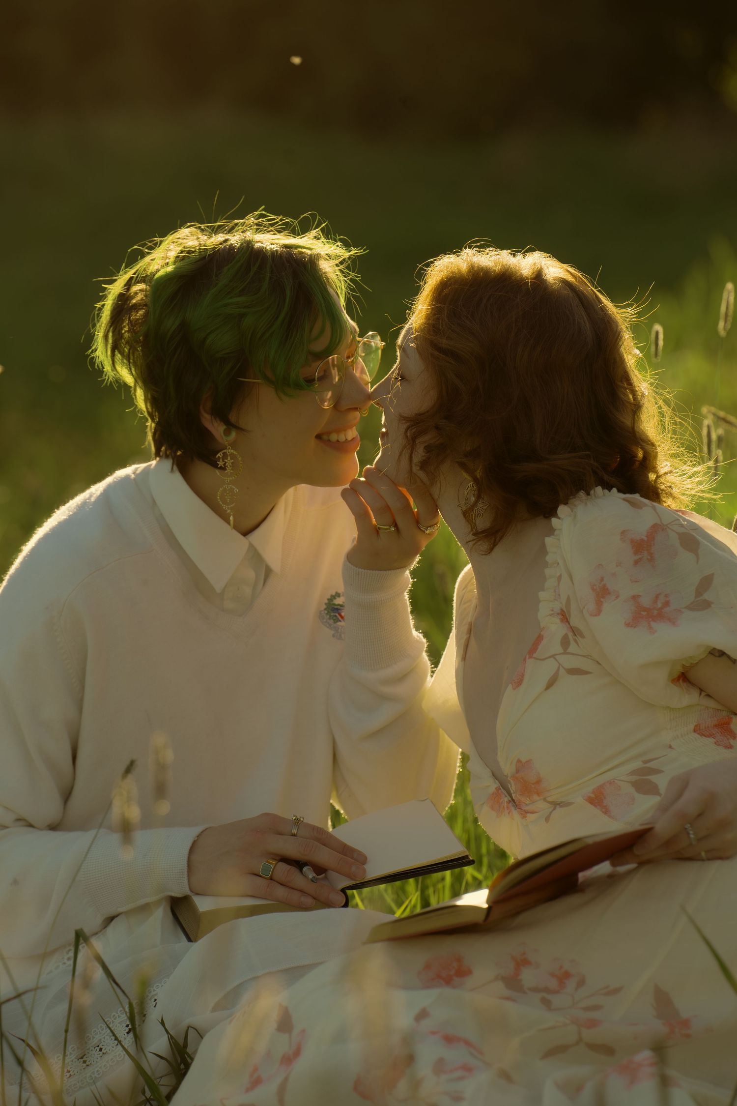 Two people share a romantic moment in golden sunlight, sitting in grass with a book, one with green-tipped hair.