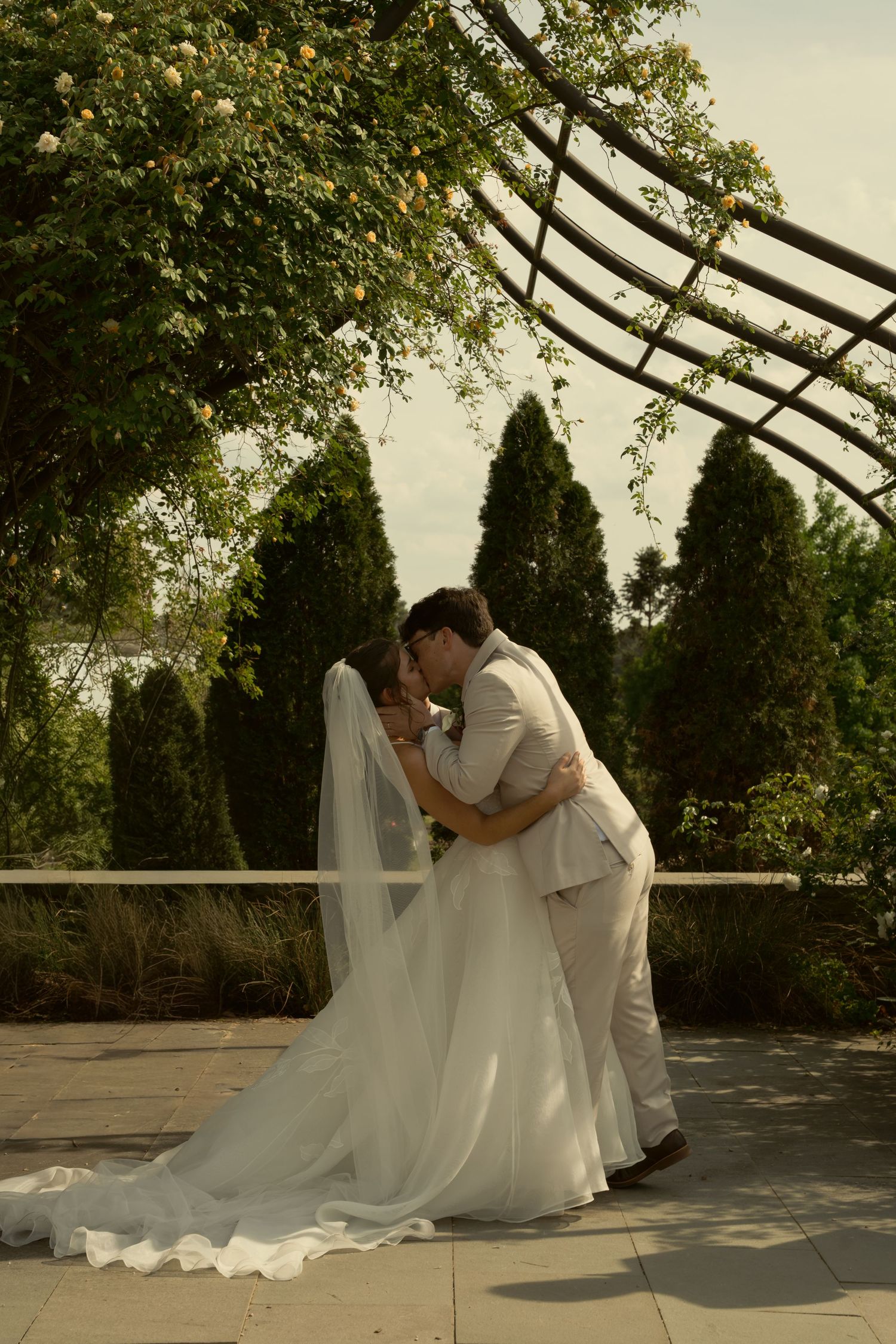 Bride and groom share a romantic dip kiss under a floral garden arbor, surrounded by lush greenery.