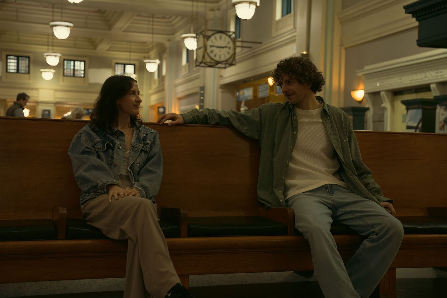 People sit on wooden benches in an indoor space with vintage lighting fixtures.