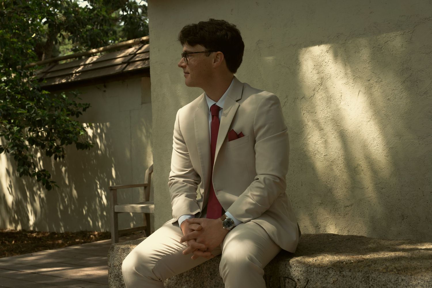 Groom in tan suit and red tie sits outdoors in dappled sunlight during wedding day portraits.