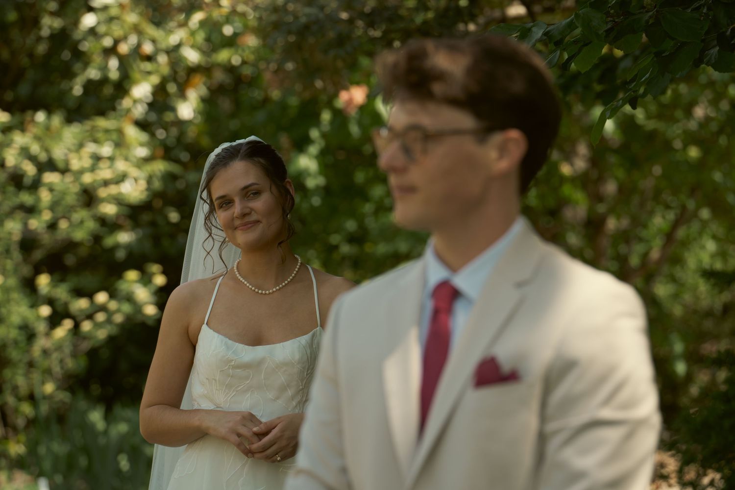 Bride in white gown and veil smiles at groom in beige suit with red tie during outdoor wedding ceremony.