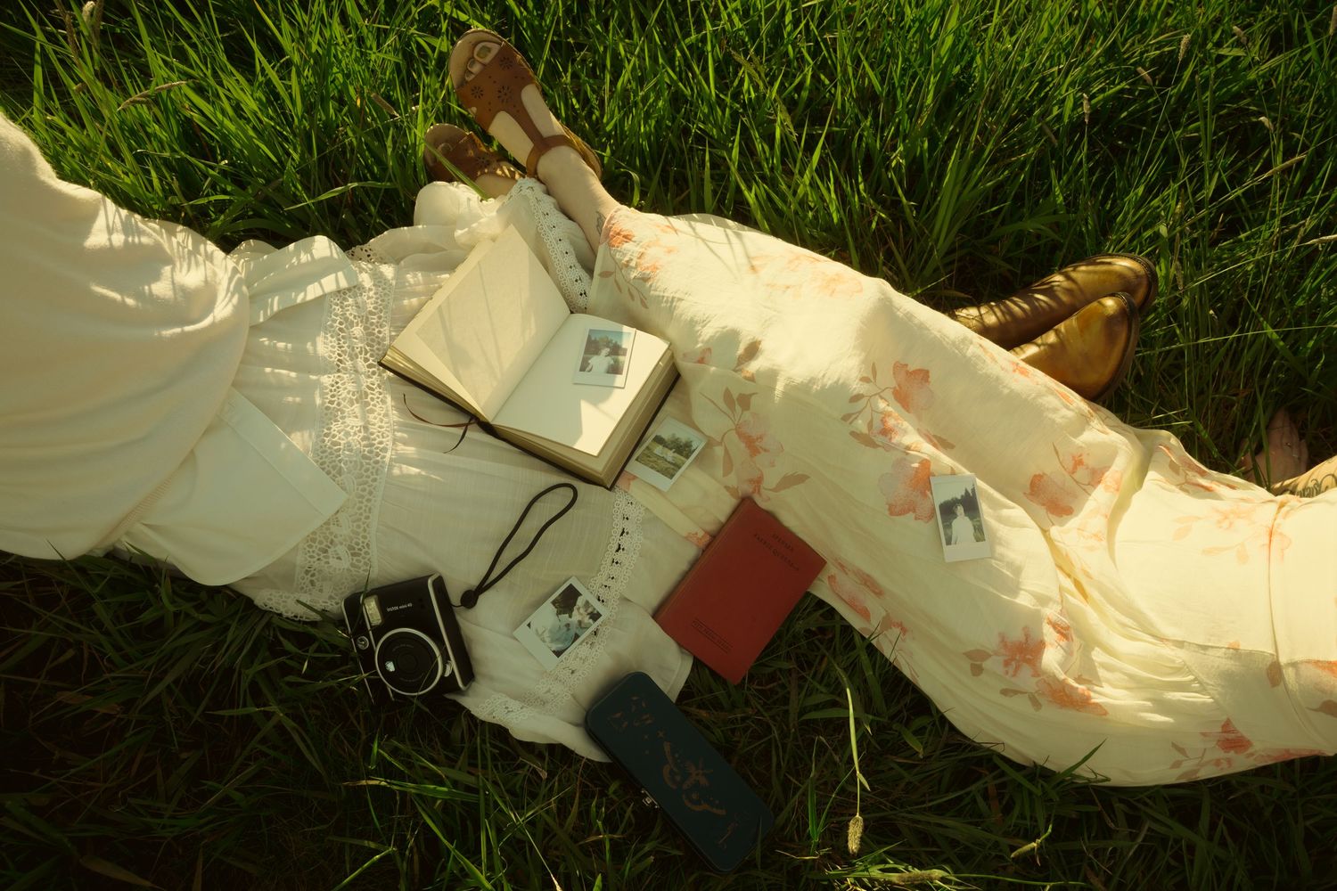 Woman in floral dress lying on grass with camera, book, and red journal beside her legs in golden sunlight.