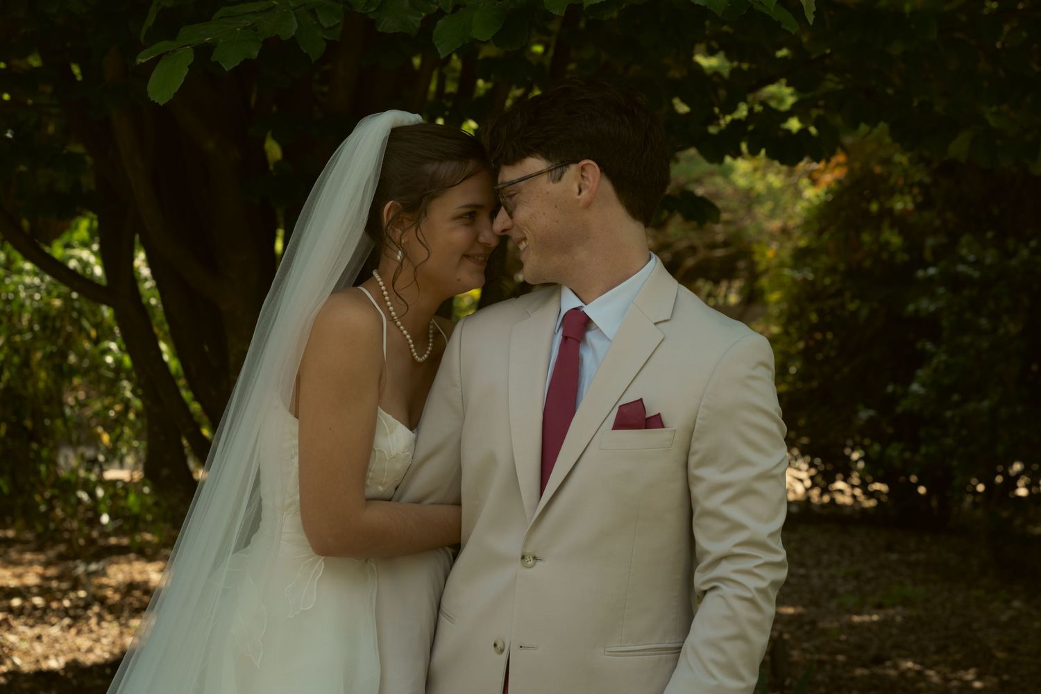 Bride and groom in tan suit share a romantic forehead touch in a garden setting.