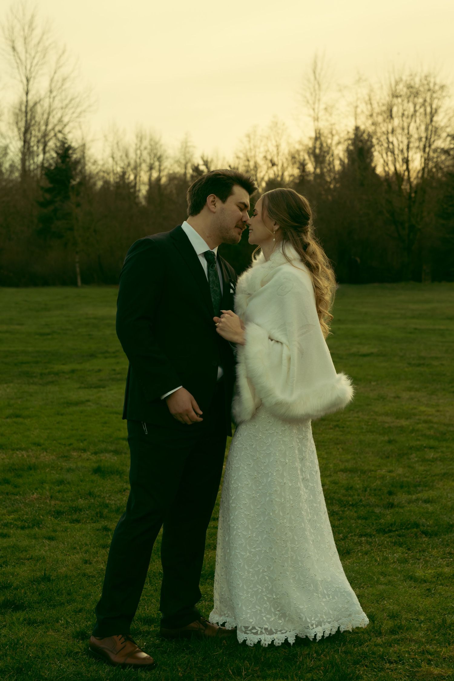 A romantic wedding couple embraces in a grassy field at dusk wearing a lace dress and dark suit.