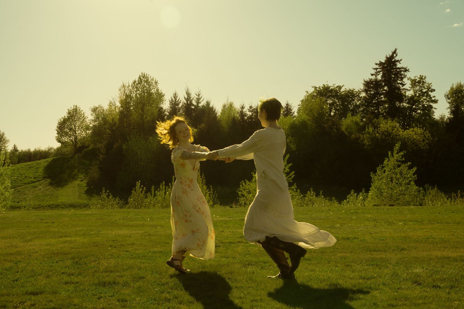 Couple in white dresses spinning and dancing joyfully in a sunny green field.