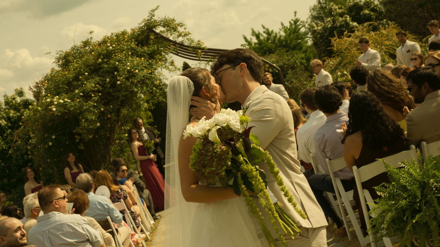 Bride and groom share a kiss at an outdoor wedding ceremony surrounded by guests and a floral arch.