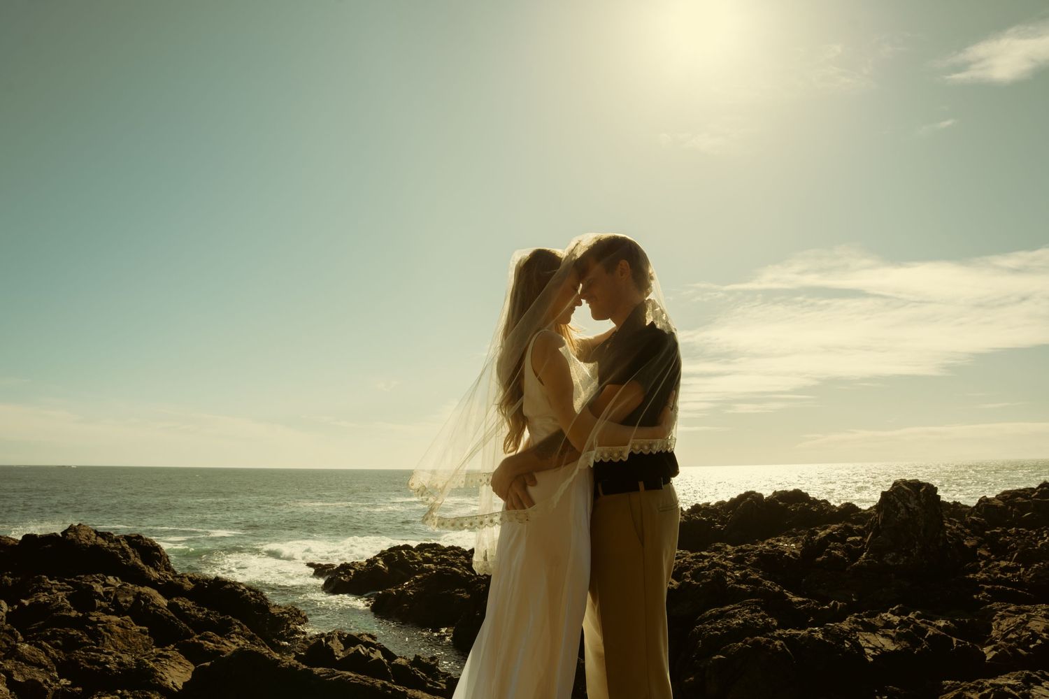 Bride and groom share a romantic kiss on rocky oceanside cliffs with sunlit waves and blue sky backdrop.