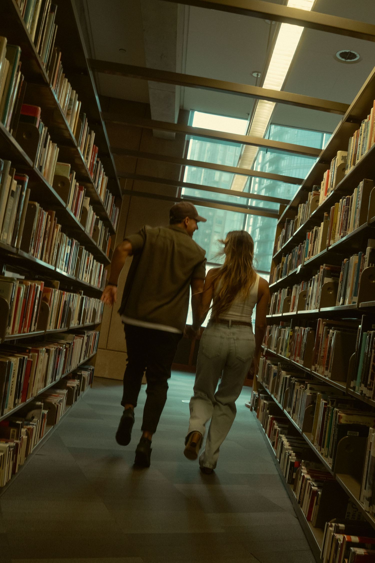 People walking between tall library bookshelves with fluorescent lighting overhead in a quiet study environment.