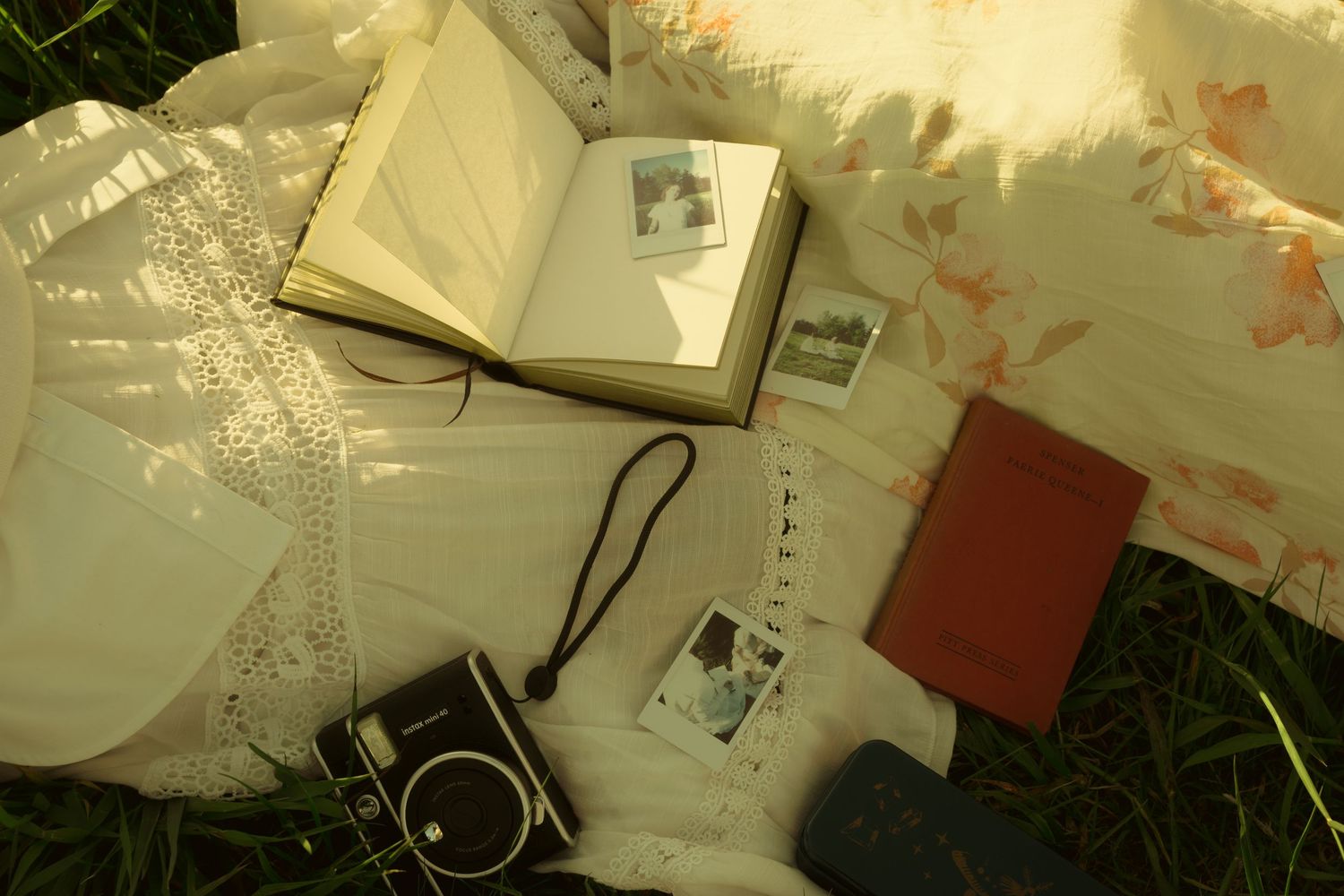Vintage film camera, open journal, and Polaroid photos arranged on a floral blanket in soft light.