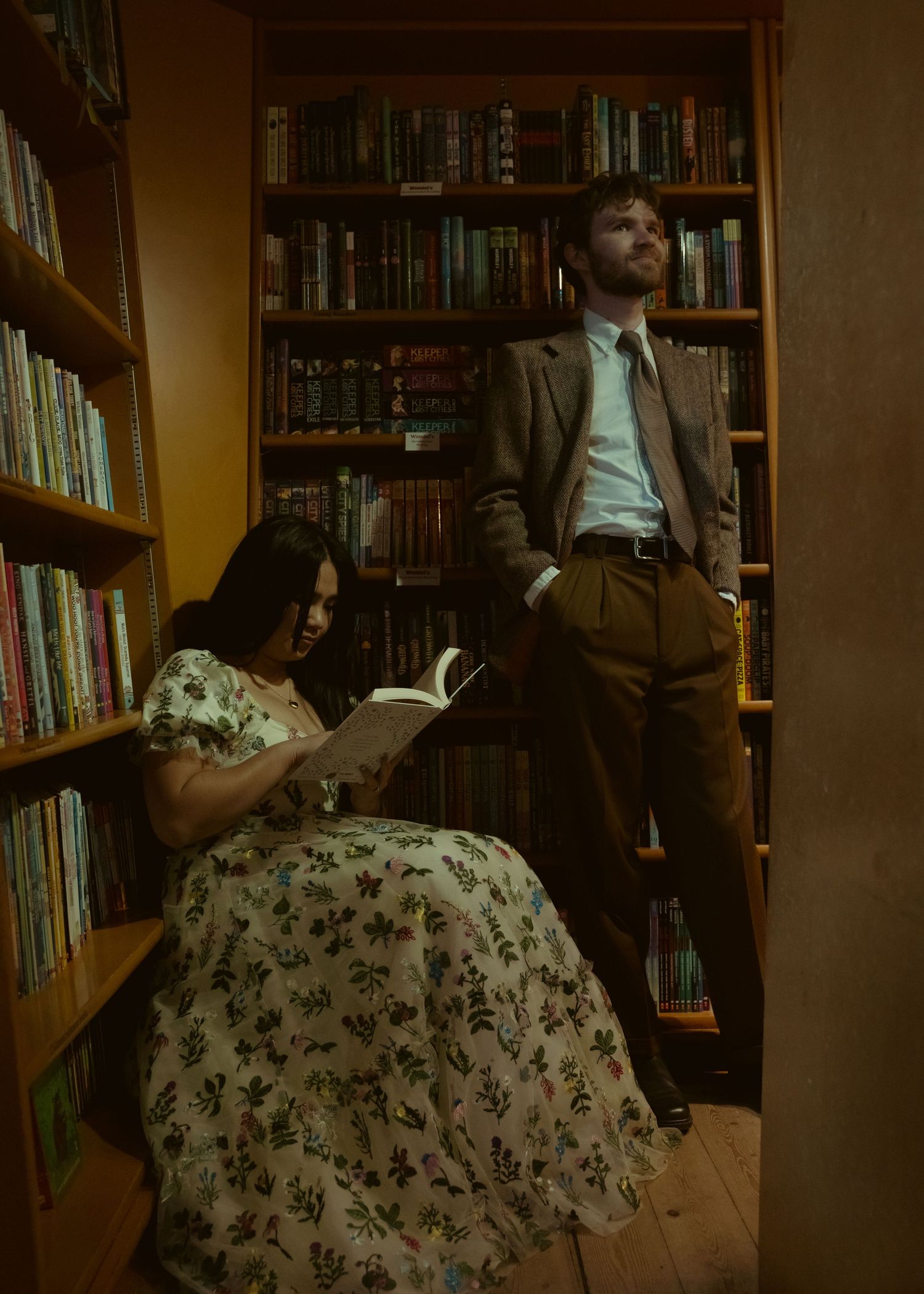 A woman in a floral dress reads a book in a cozy library while a man in a suit stands gazing upward at bookshelves.