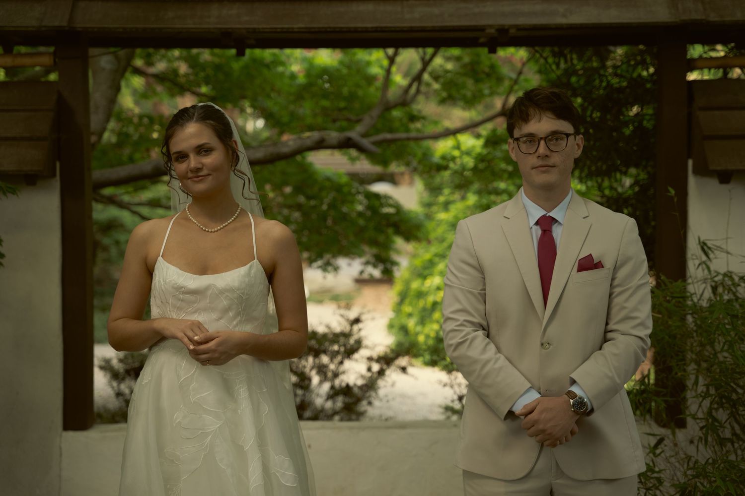 Bride in white dress and groom in tan suit standing under garden pergola.