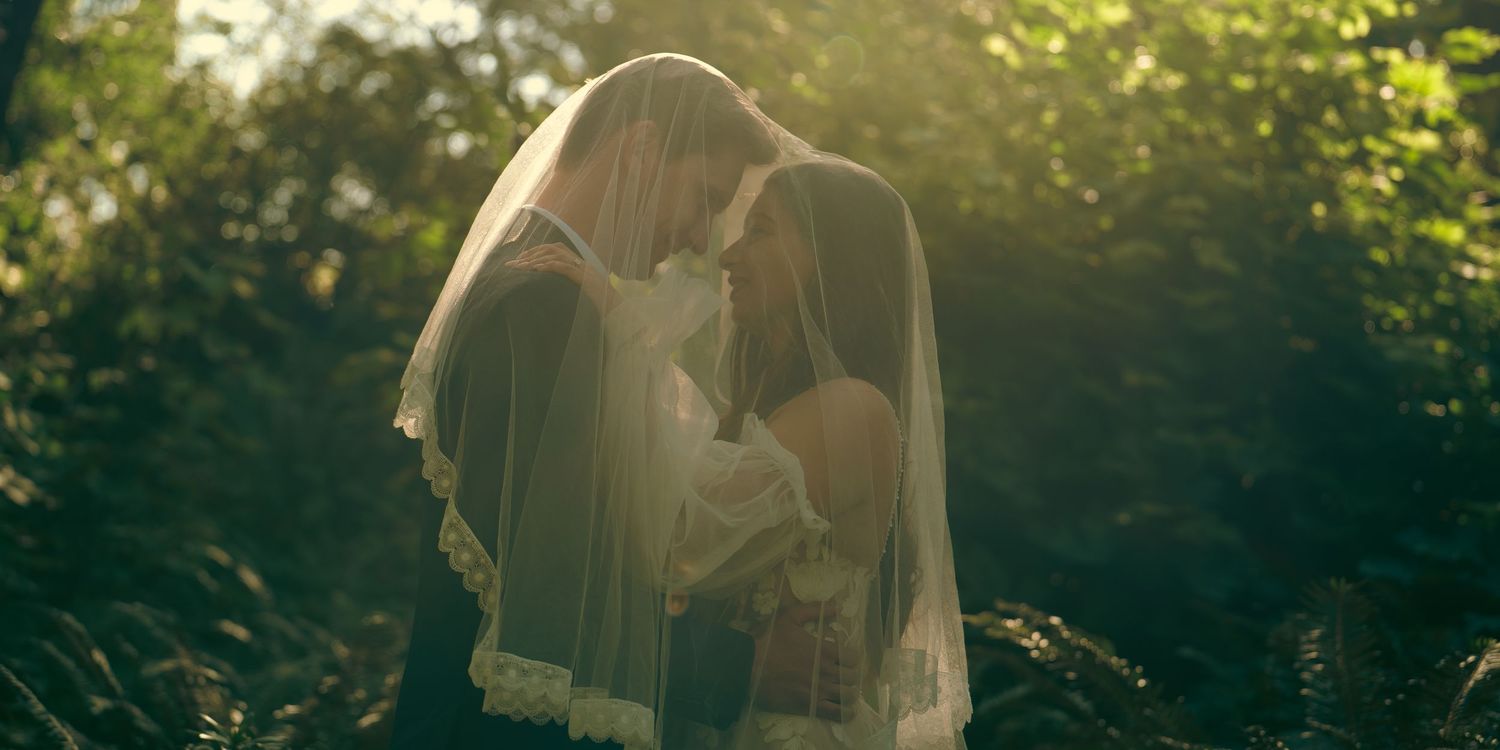 Bride and groom sharing romantic moment under wedding veil in sunlit forest setting.