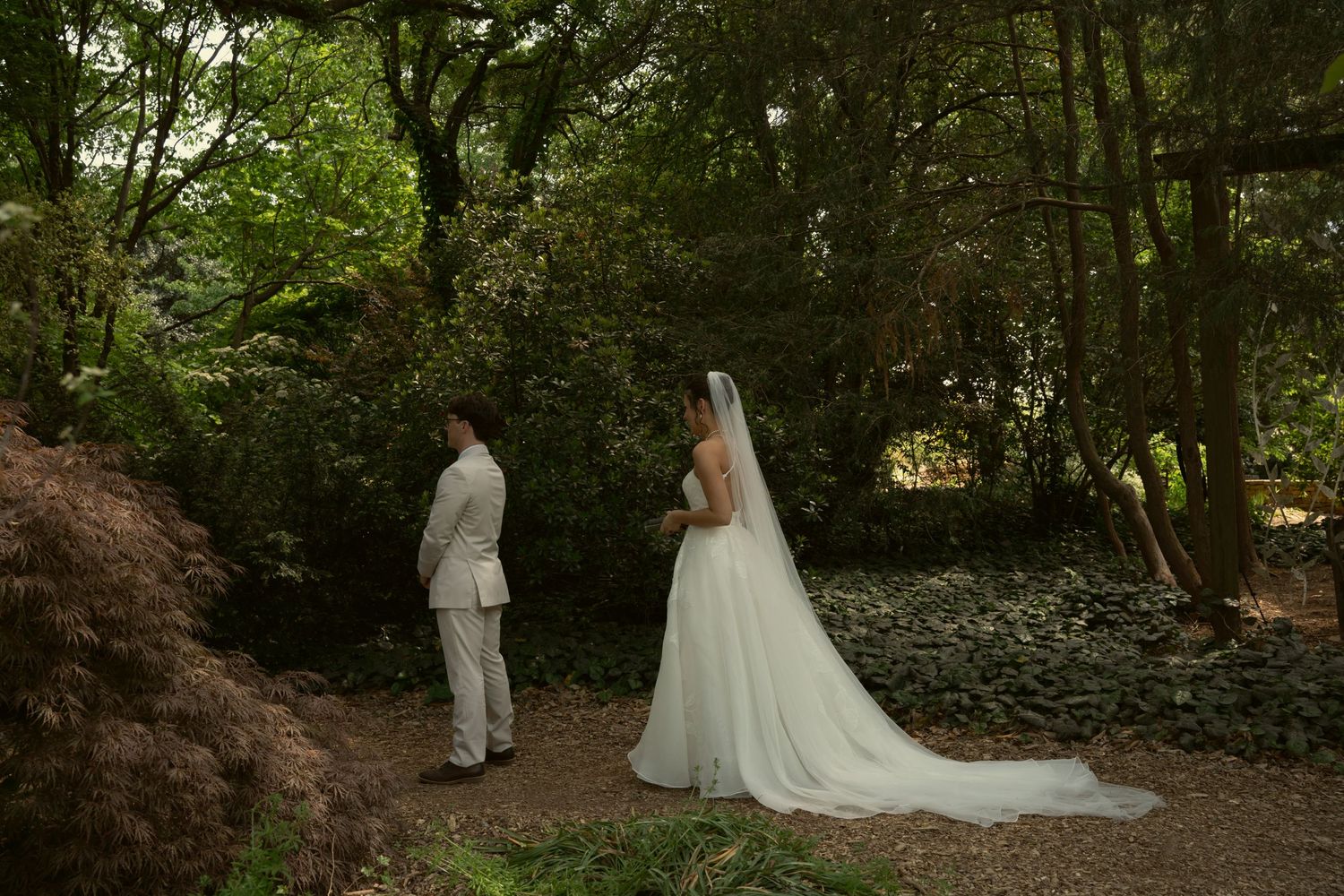 Bride and groom stand together on garden path surrounded by lush green trees during wedding photo session.