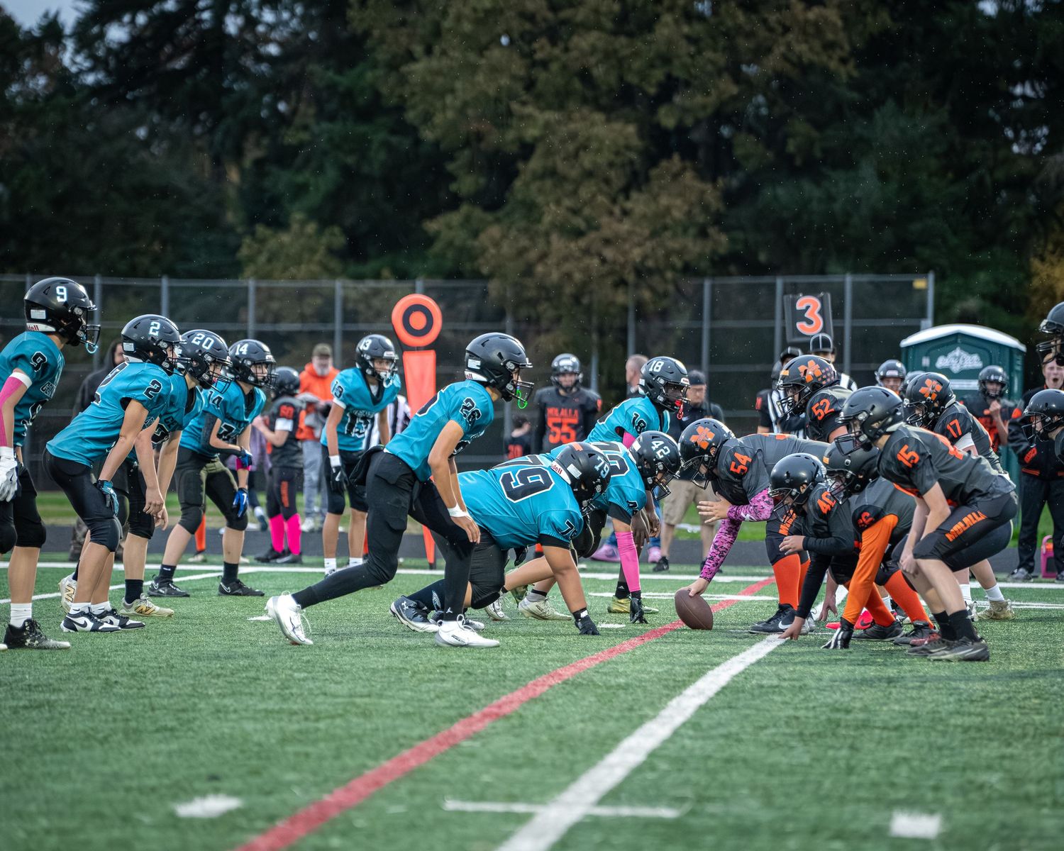 Football team practices in formation on turf field with officials and spectators visible.