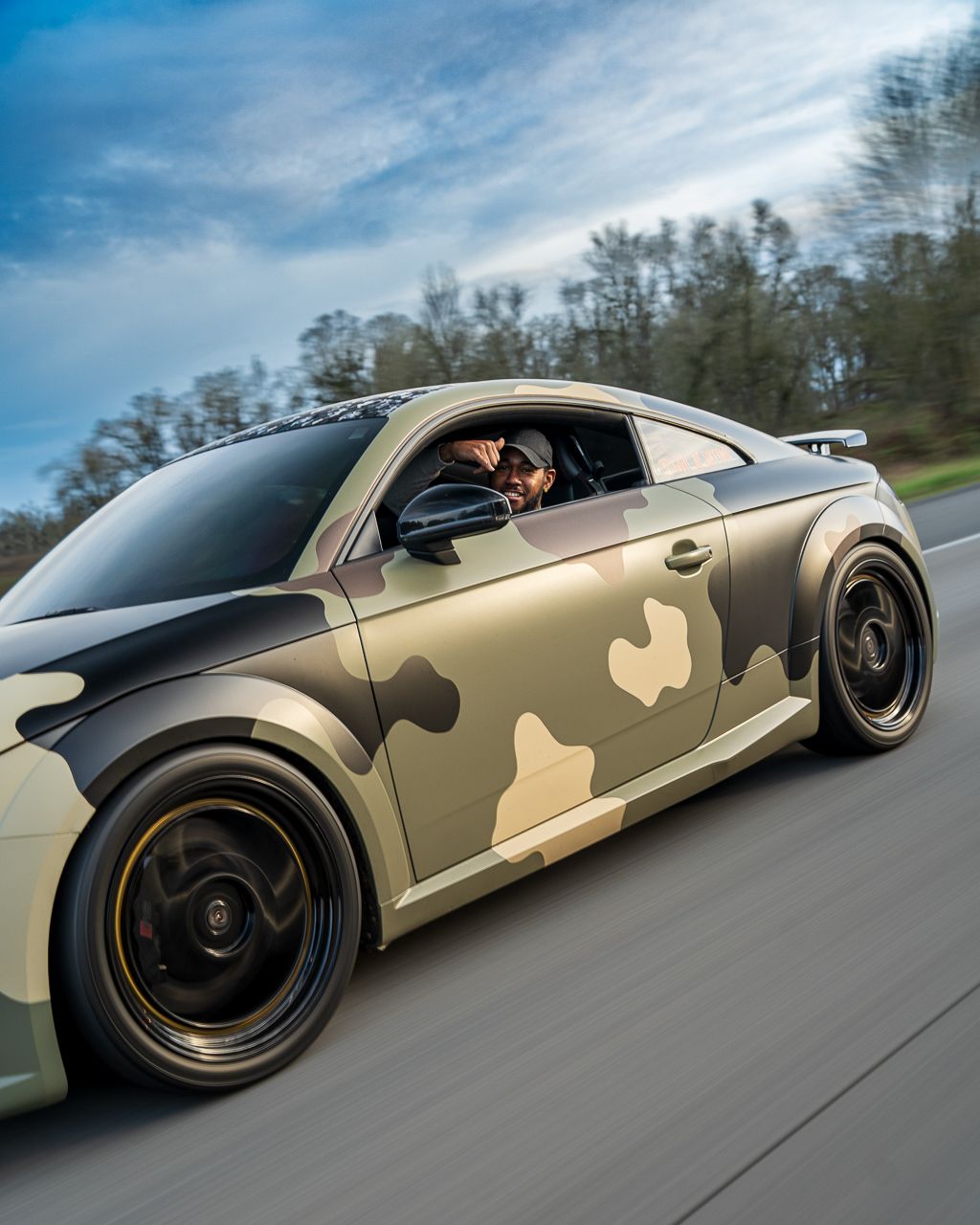 Camouflage-wrapped sports car photographed in motion on an open road.