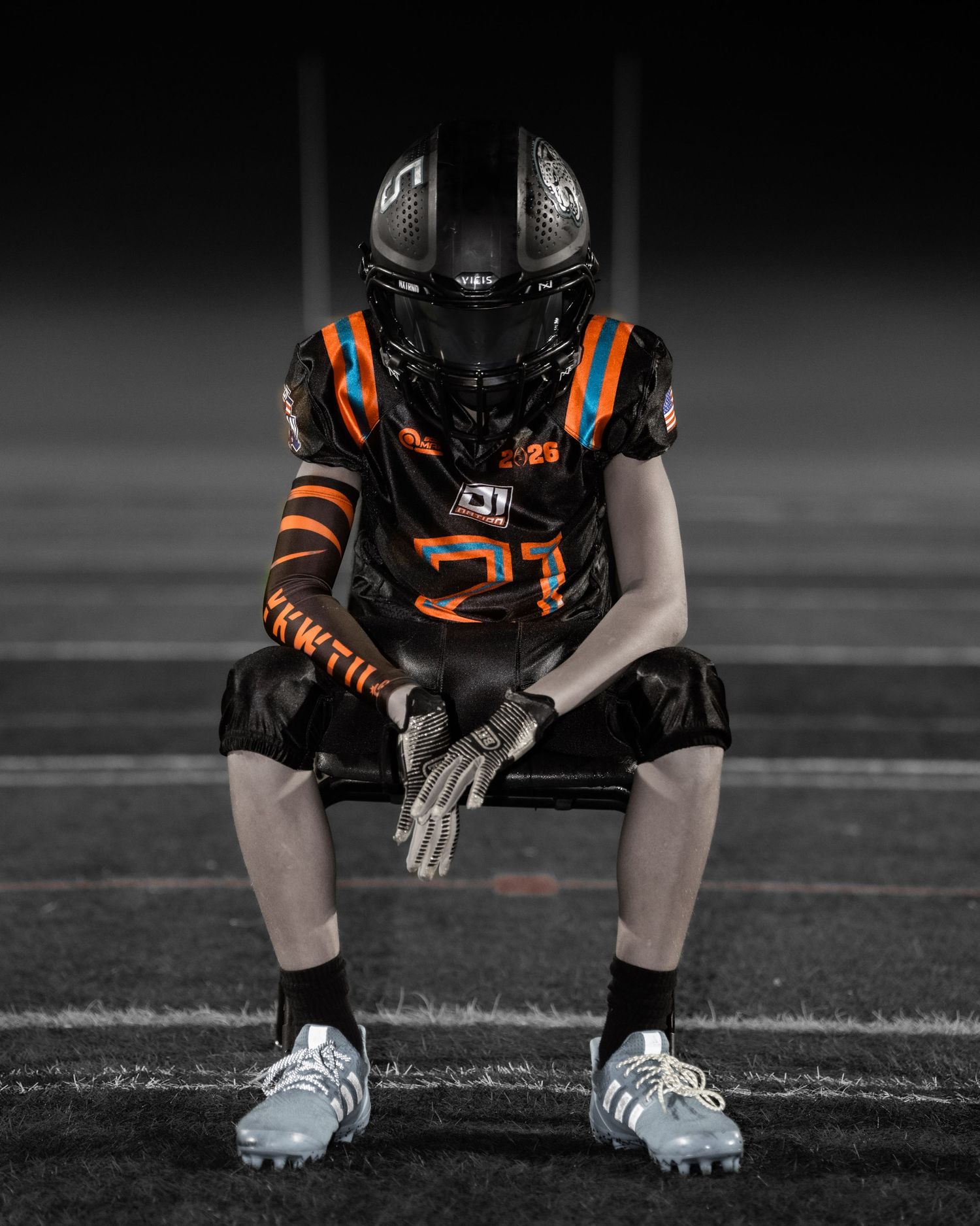 Football player in black and orange uniform sits on field wearing silver cleats in selective color sports photograph.