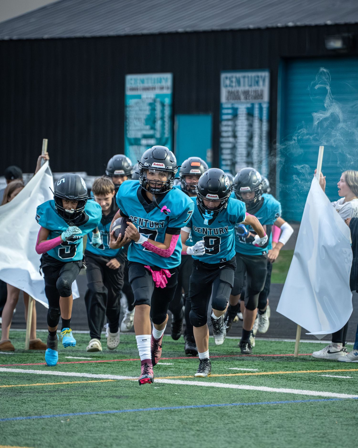 Football team in teal uniforms runs through banner entrance before game.