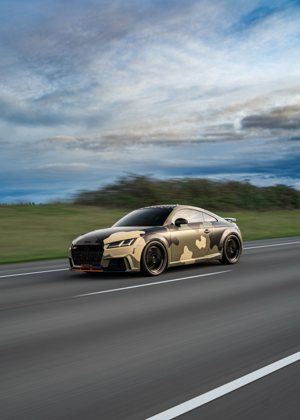Gold sports car with custom body kit speeding down highway under cloudy skies.