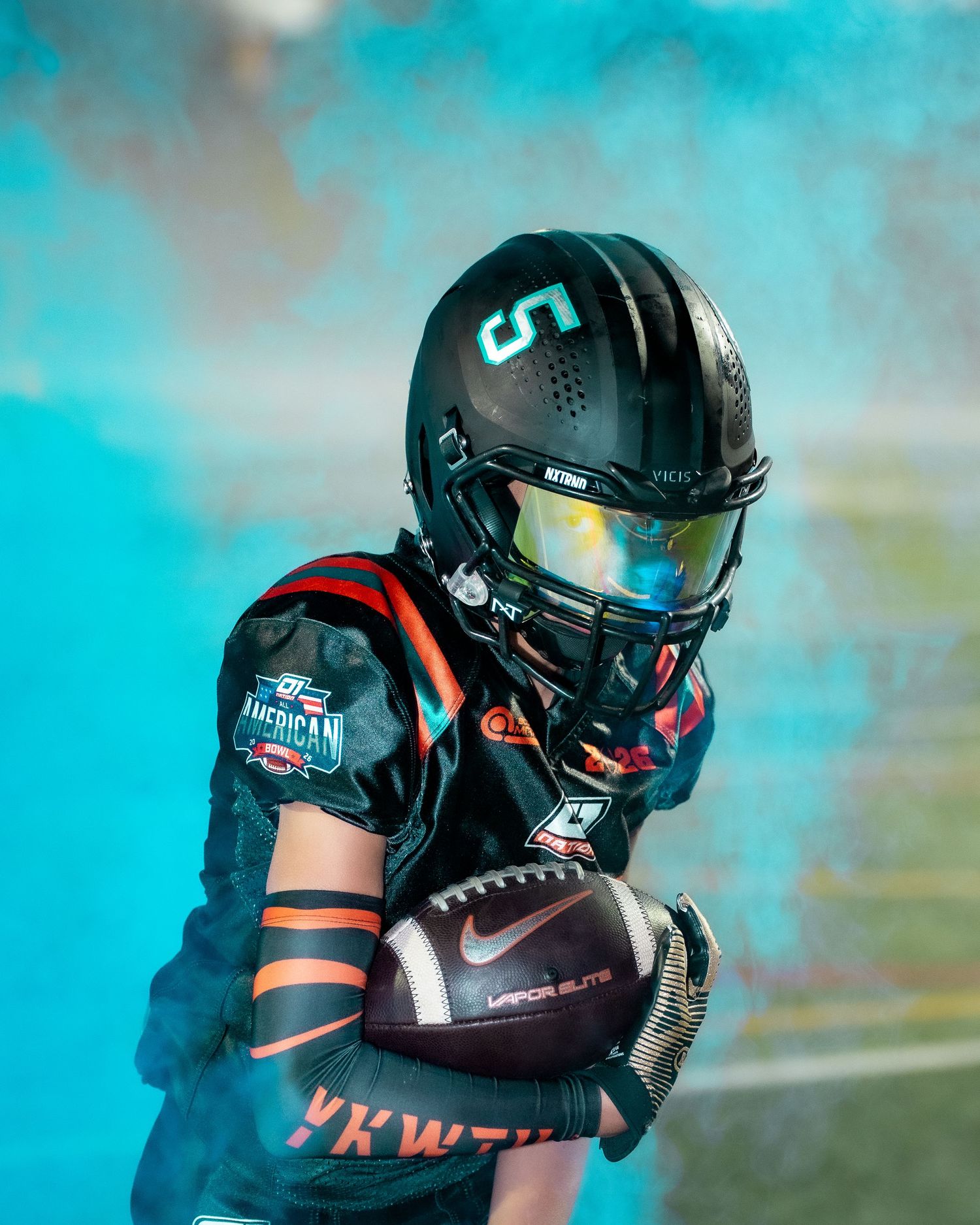 Football player in black helmet and uniform looking to the side against turquoise background.