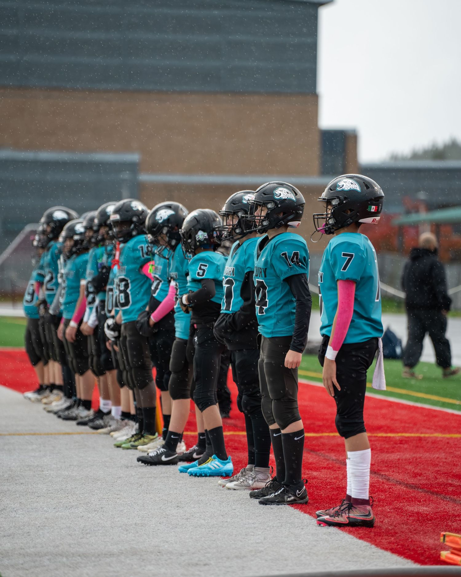 Football team in teal jerseys lines up on sideline during game.