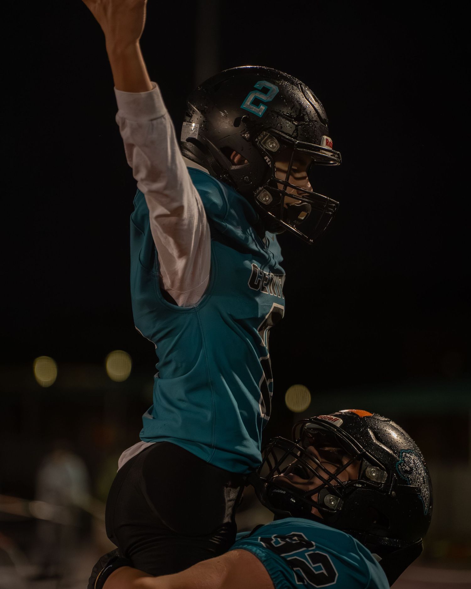 Football player in teal jersey and black pants crouches in ready position during nighttime game.
