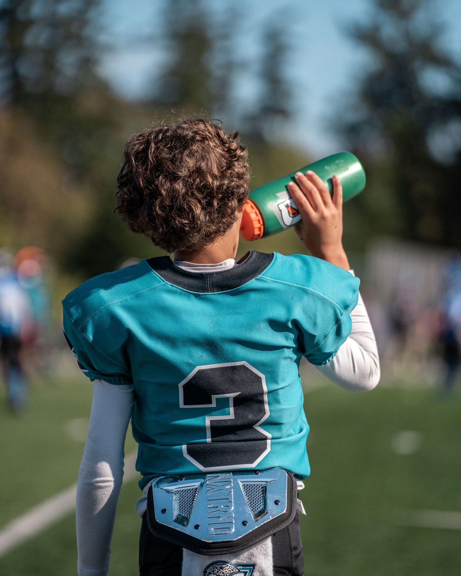 Football player in teal jersey drinking from water bottle on sunny field.