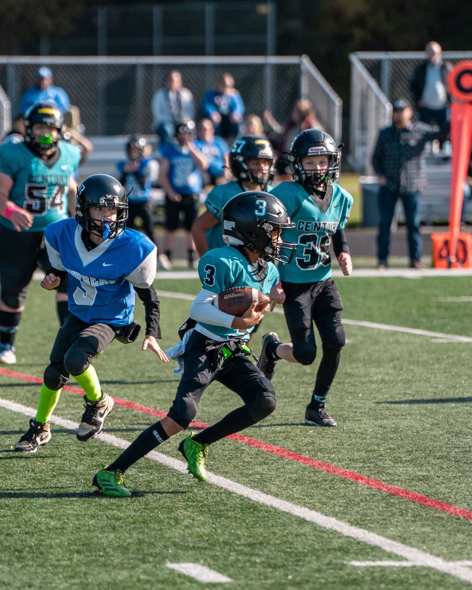 Youth football players in teal and white uniforms run plays during a game.