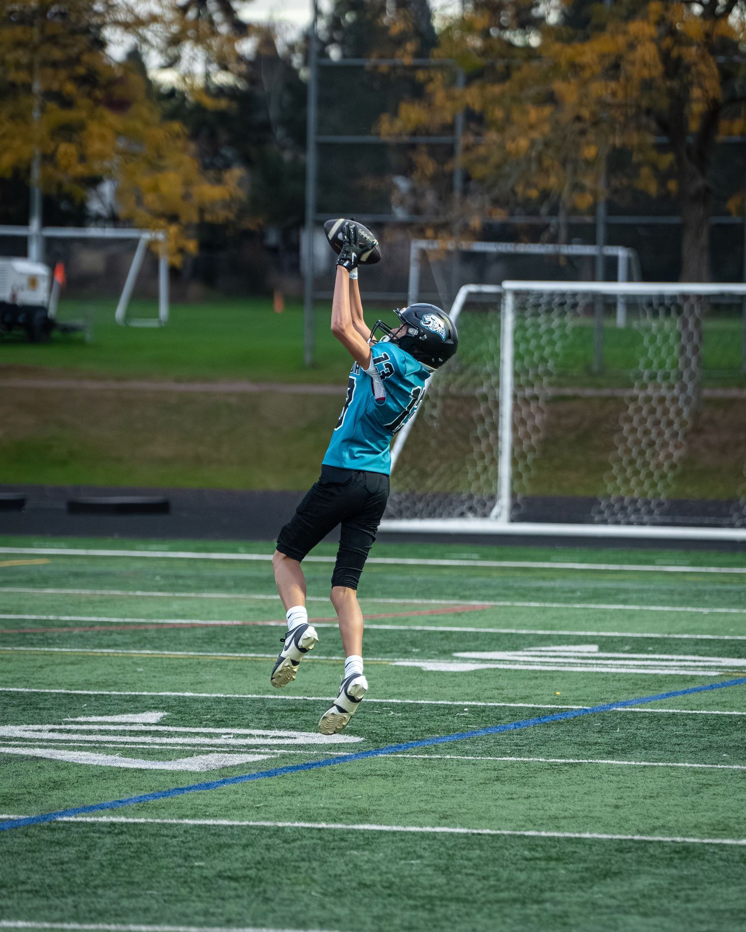 Football player practices kicking technique on sports field with goal posts visible in background.