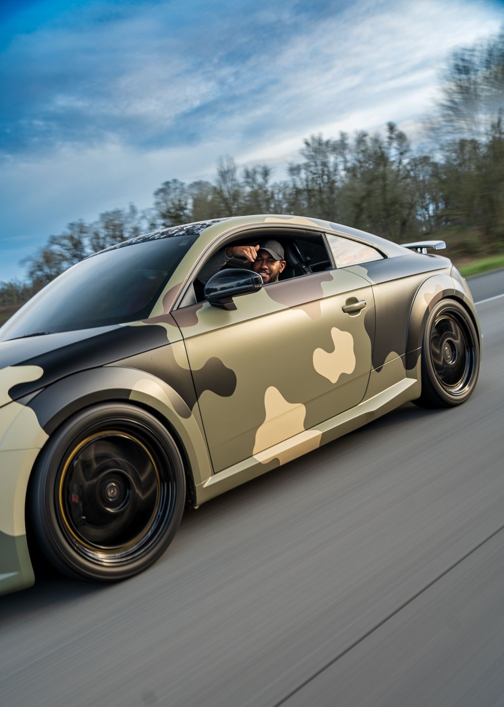 Custom camouflage-wrapped luxury car photographed in motion on open road.