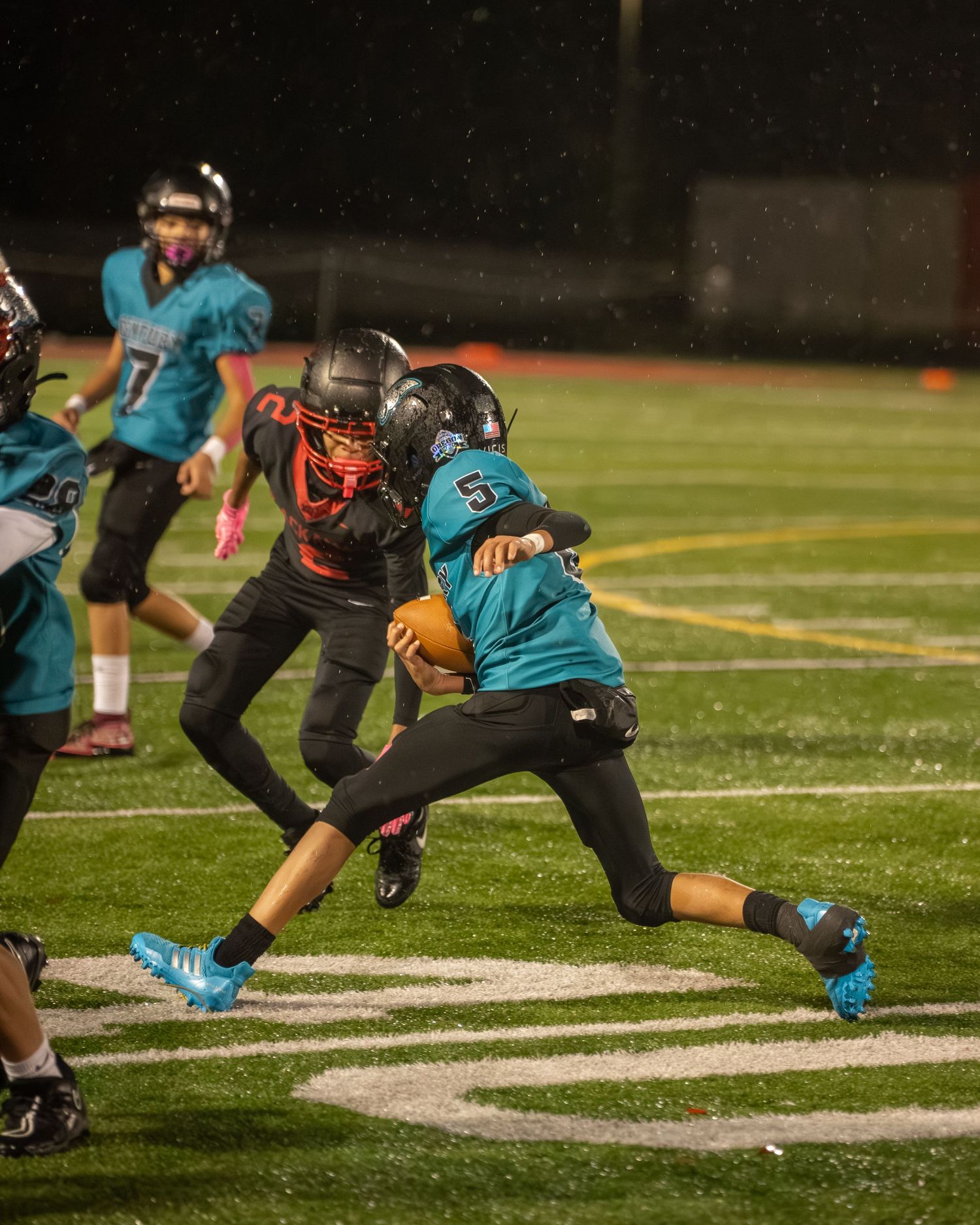 Football players in teal and black uniforms engage in gameplay near scrimmage line.