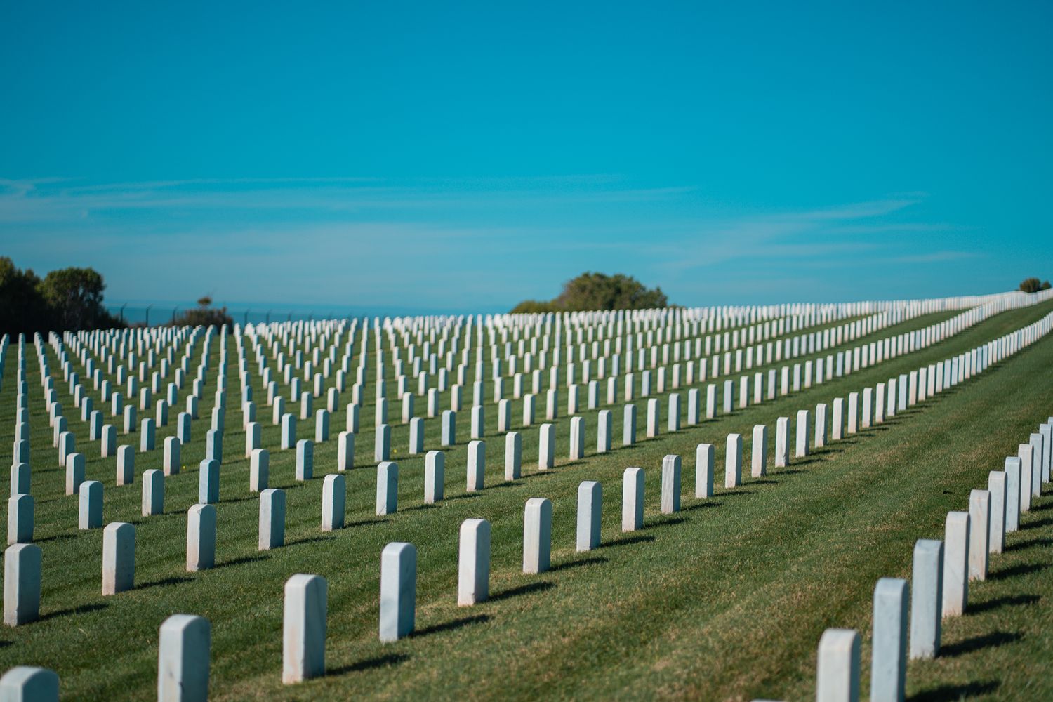 Rows of white military headstones stretch across a memorial cemetery under bright blue sky.