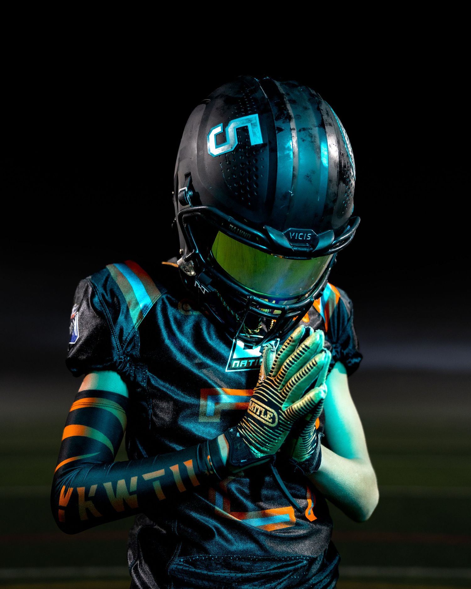 Youth football player in teal uniform and helmet with number 5 stands under dramatic lighting.