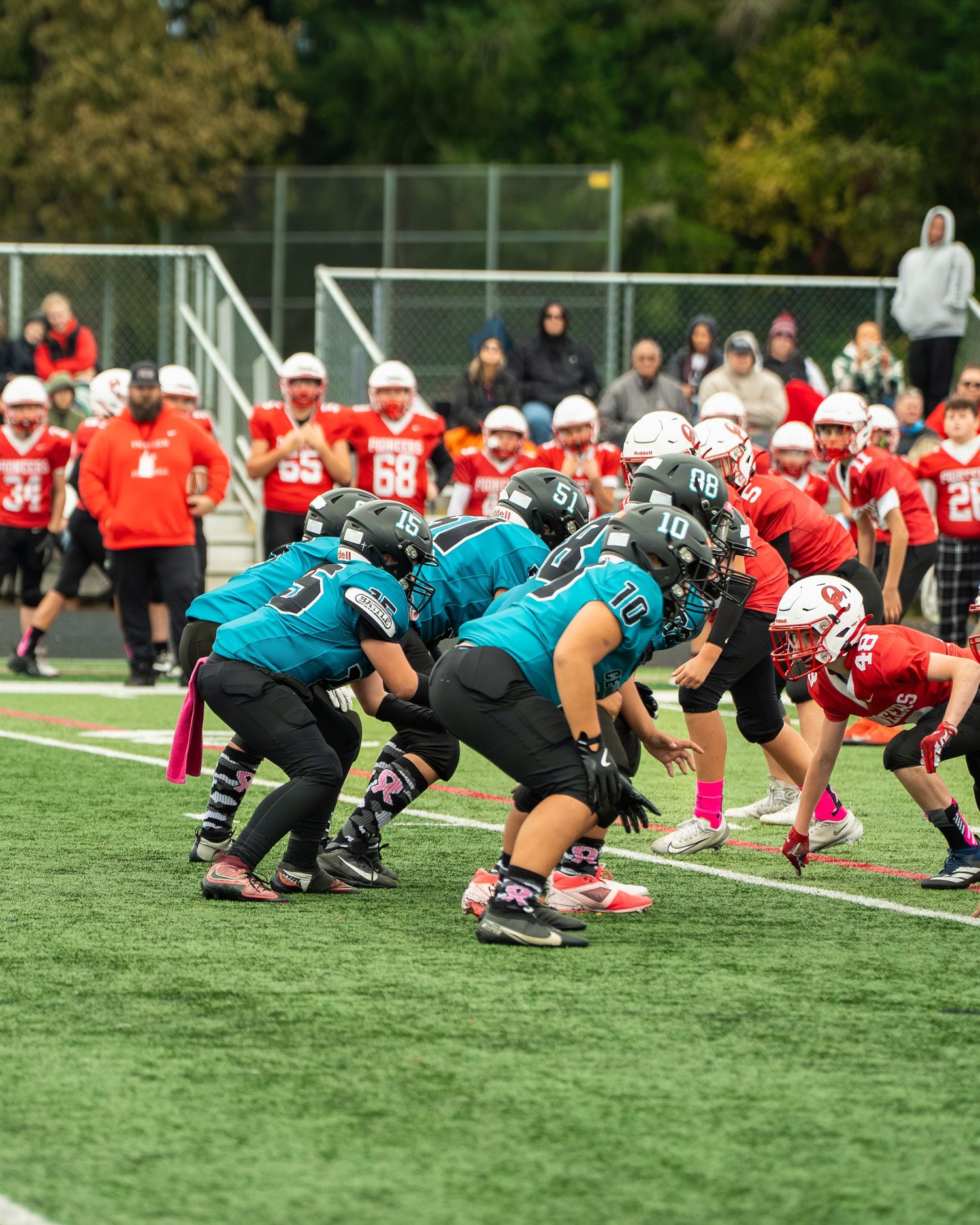 Football teams in teal and red uniforms line up at scrimmage on artificial turf field.